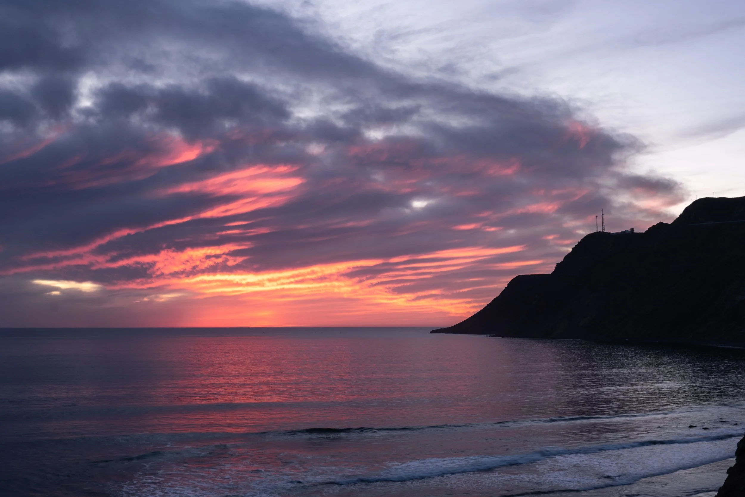 Sunset over the ocean with pink, purple, and orange clouds, and a dark silhouette of a cliff on the right side.