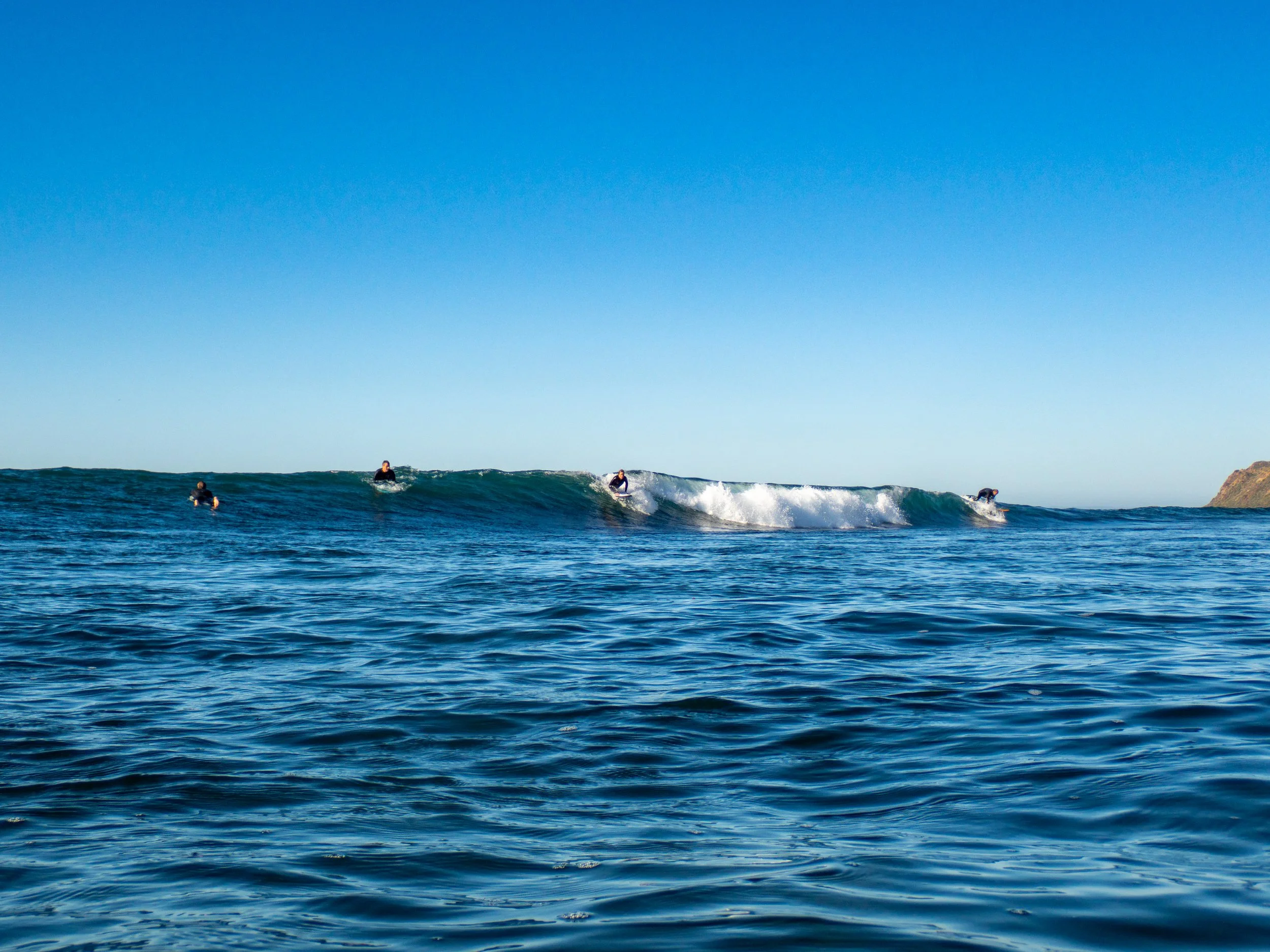 Four surfers riding or waiting for waves in the ocean on a clear day, with a blue sky and a distant coastline in the background.