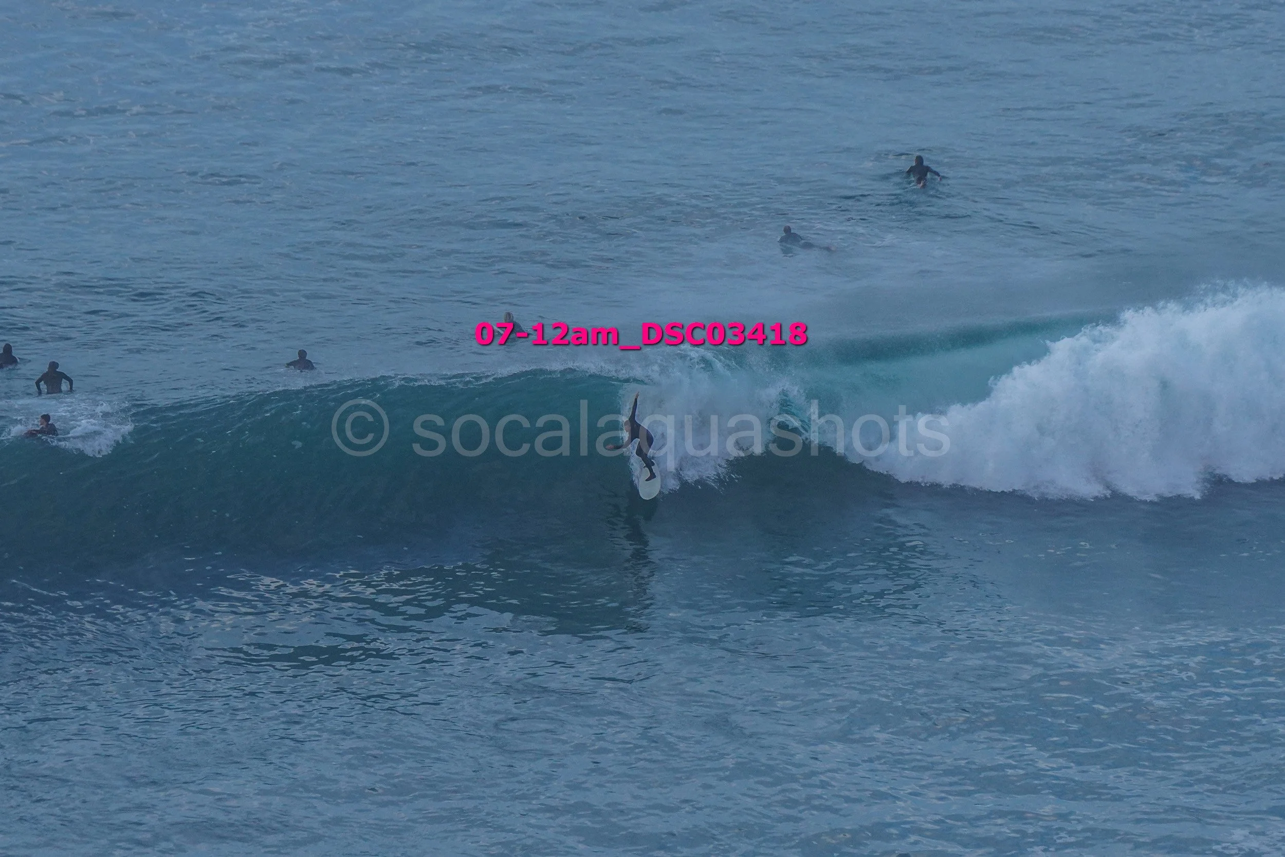 A surfer riding a large wave with several people swimming and surfing in the background in the ocean.