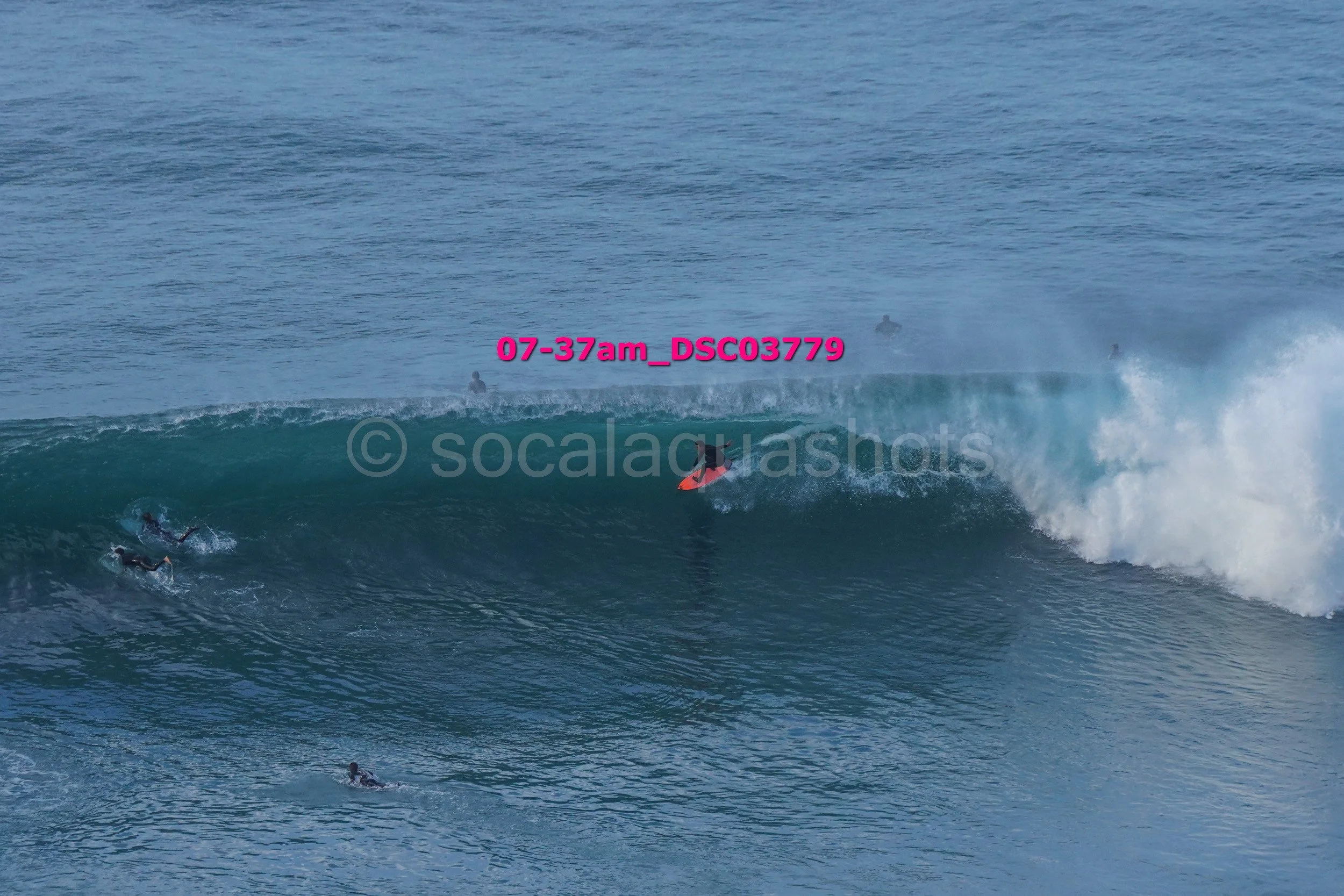 Surfer riding a large wave with other surfers waiting in the water nearby.