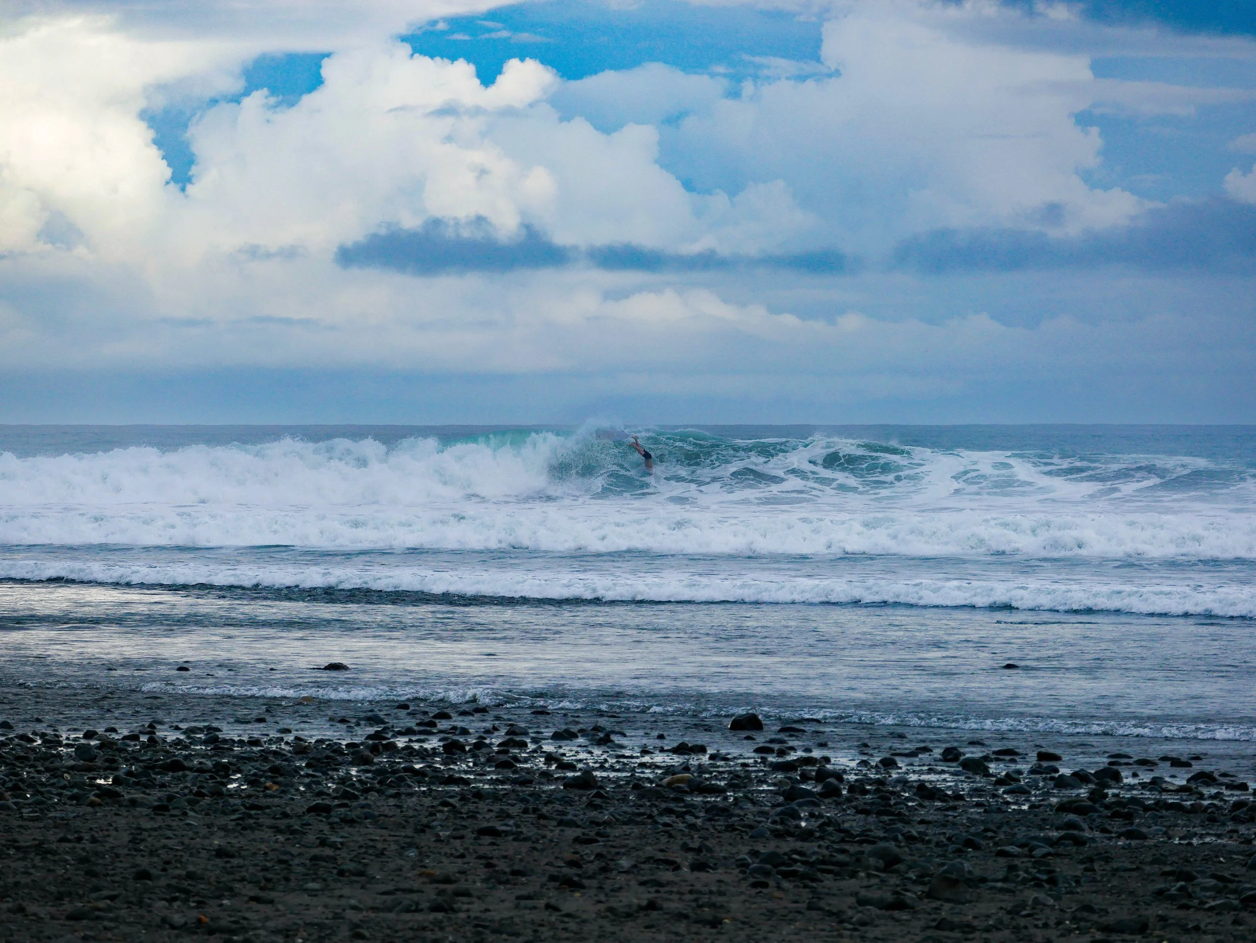 Surfer riding a wave at a rocky beach under cloudy sky