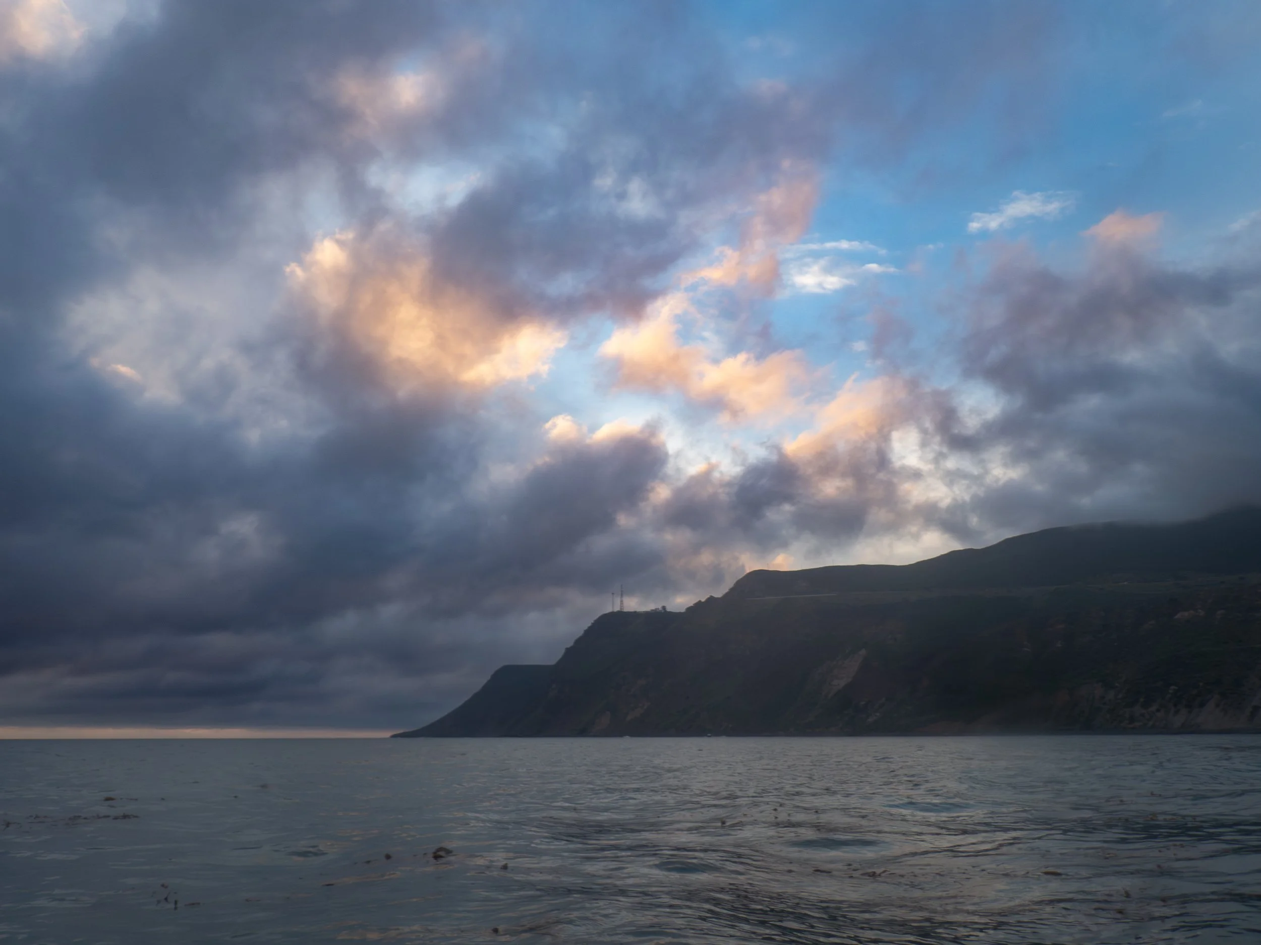 Cloudy sky over a mountain coastline during sunset with calm ocean water in the foreground.