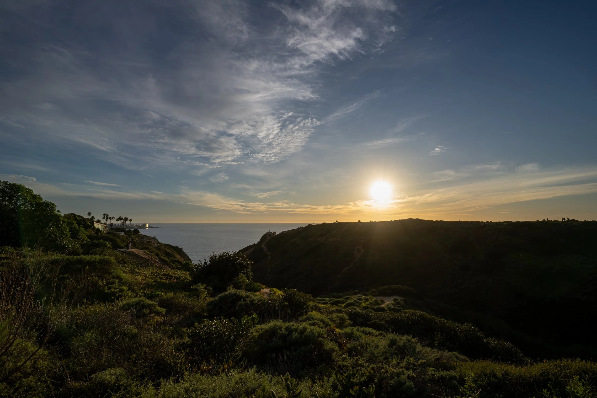 Sunset over a hillside overlooking the ocean with some palm trees in the distance and a partly cloudy sky.