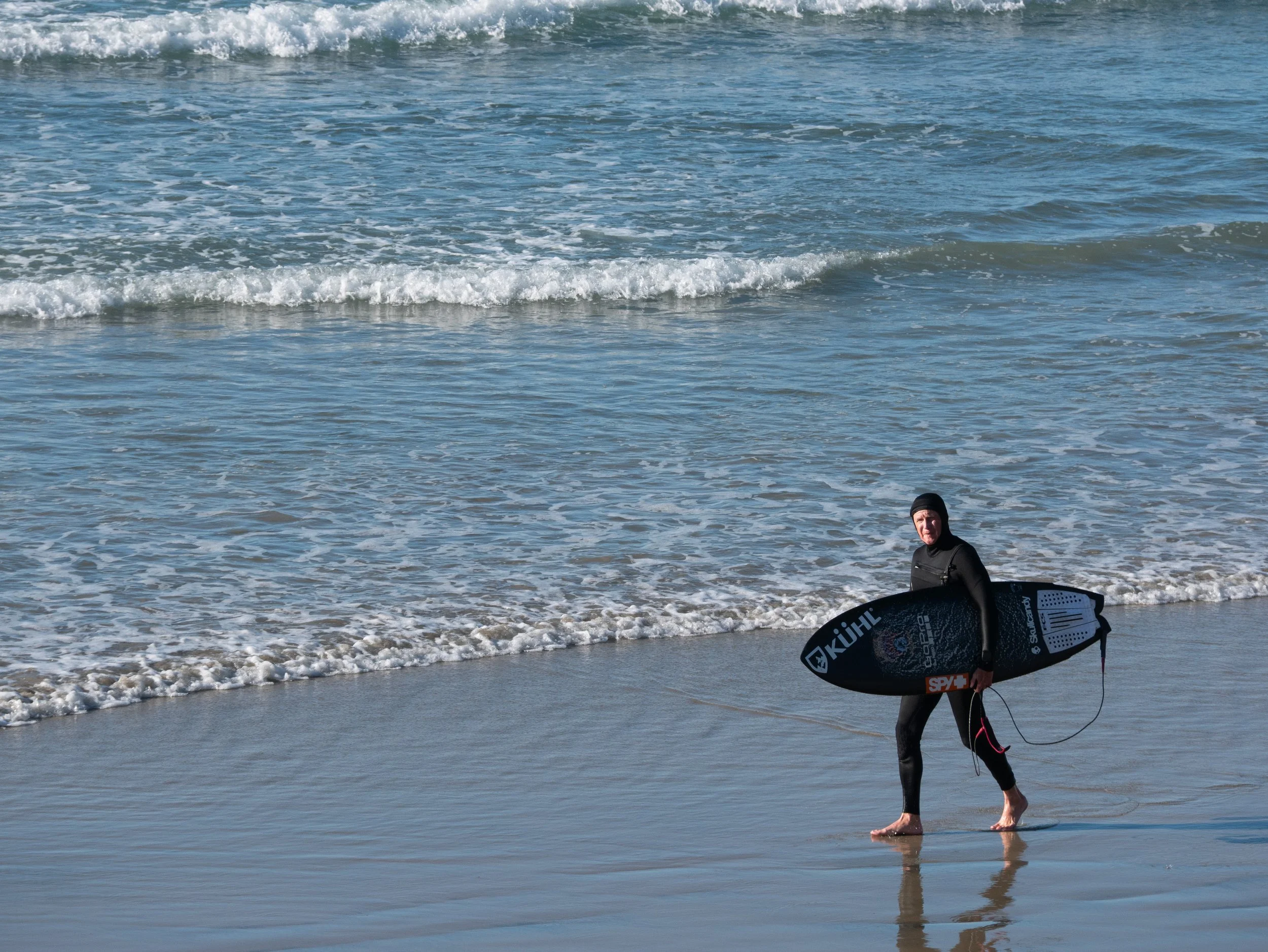A person in a wetsuit carrying a surfboard on a beach with gentle waves.