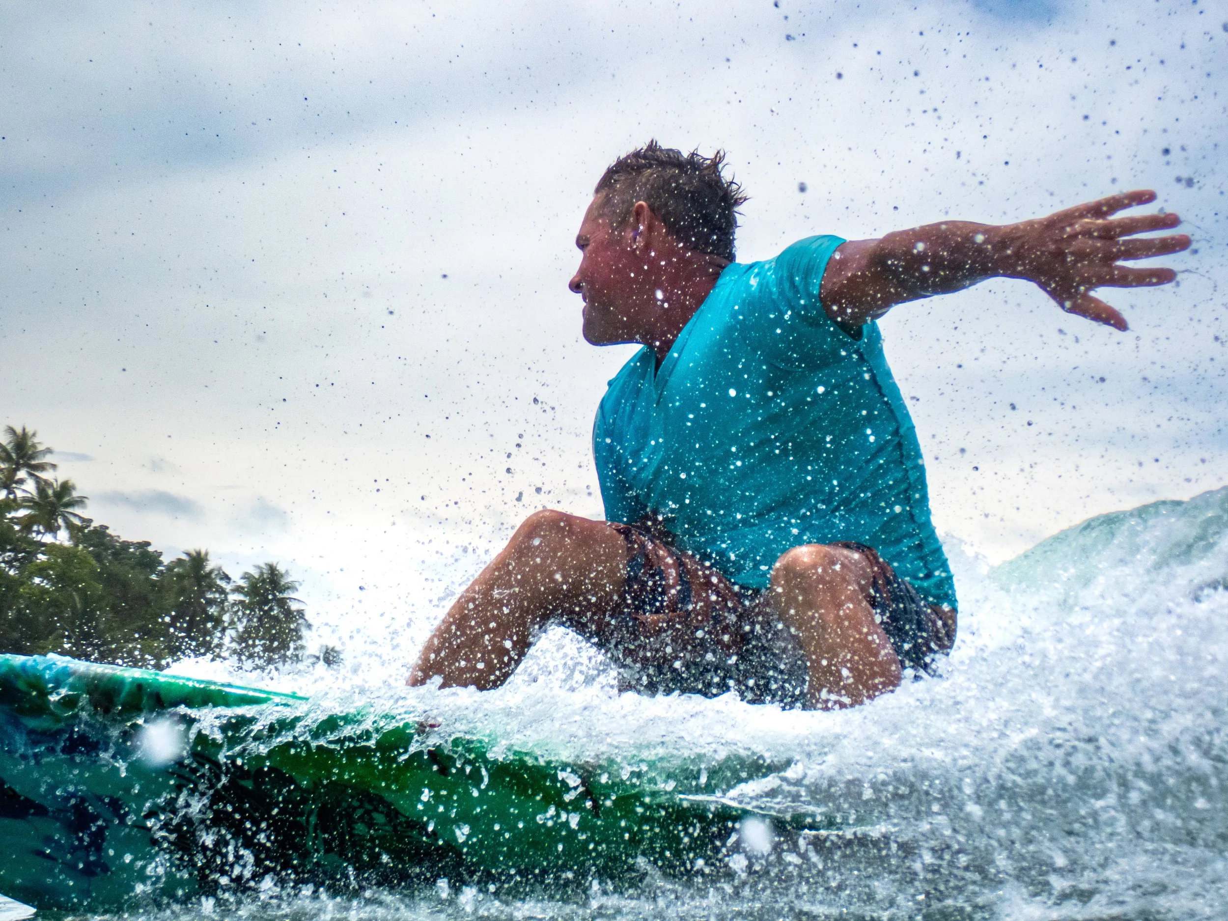 A man surfing on a green surfboard, wearing a blue shirt, kicking up water with a splash, with palm trees and a cloudy sky in the background.