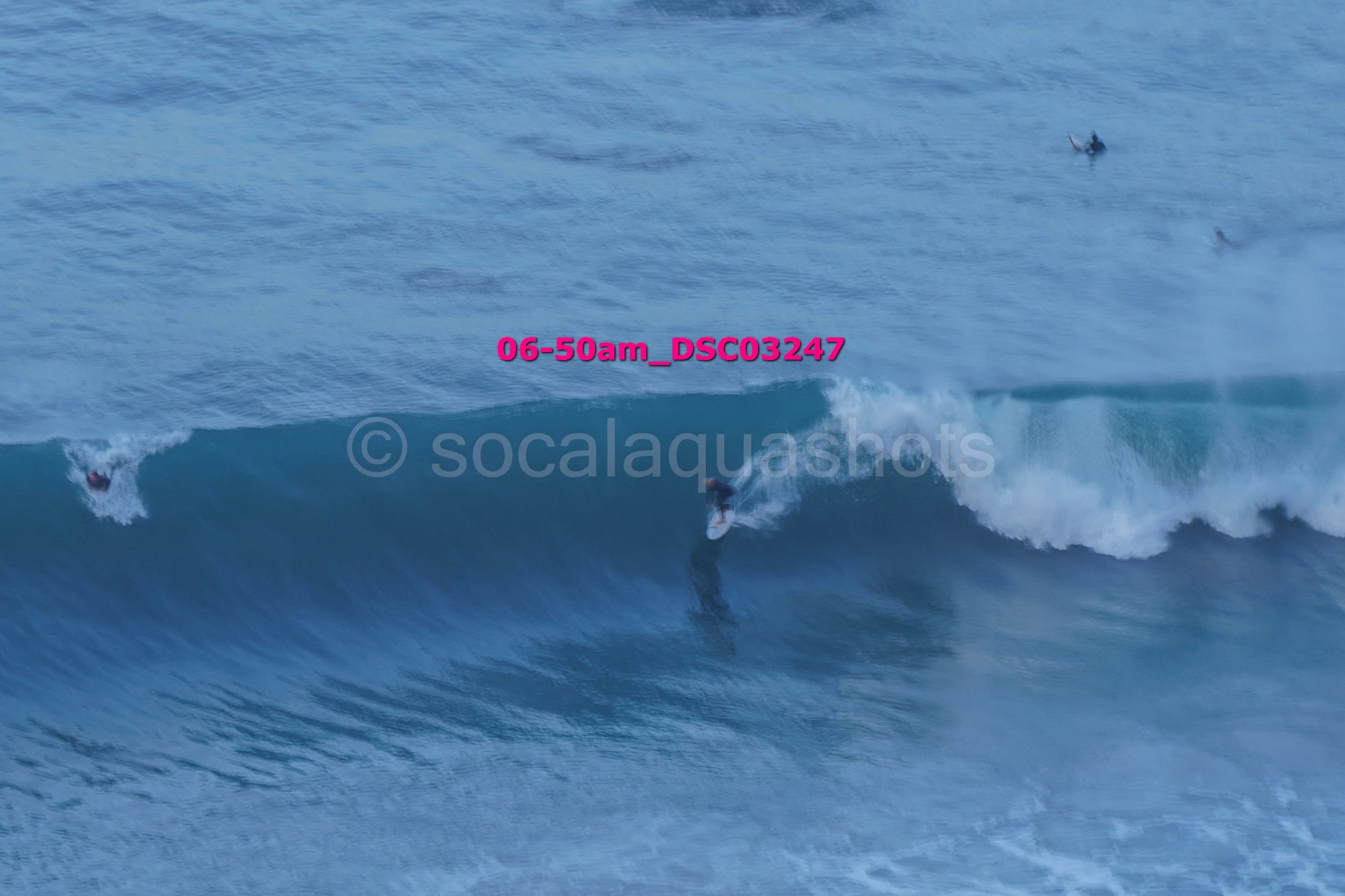 Surfers riding ocean waves at sunrise or early morning, with additional surfers paddling in the distance.