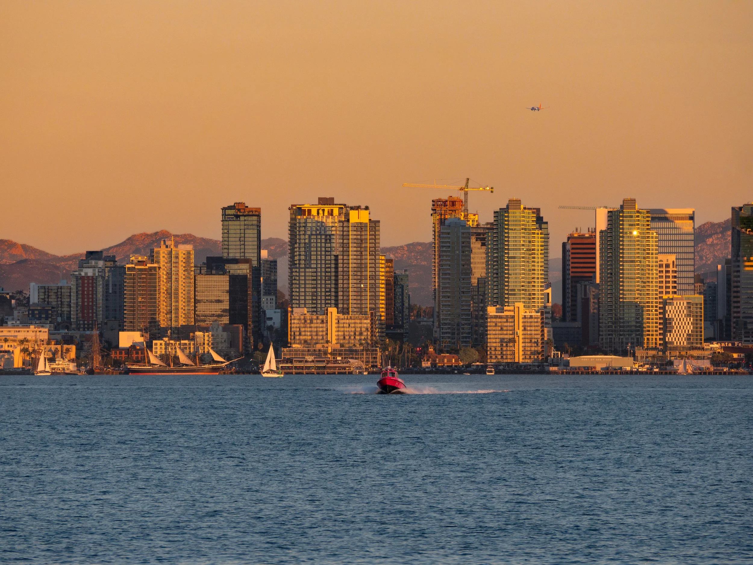 A city skyline at sunset with high-rise buildings, a boat speeding on the water, sailboats, an airplane in the sky, and a backdrop of mountains.