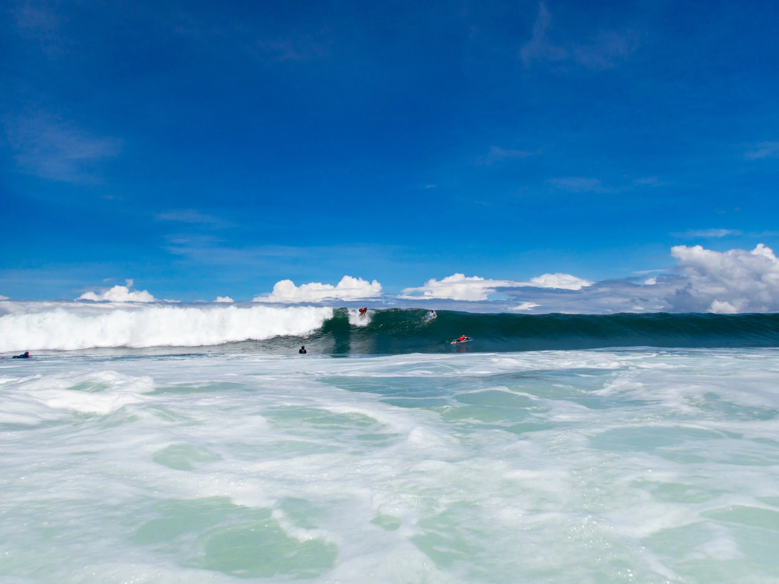 Surfers riding large ocean waves under a blue sky with scattered clouds.