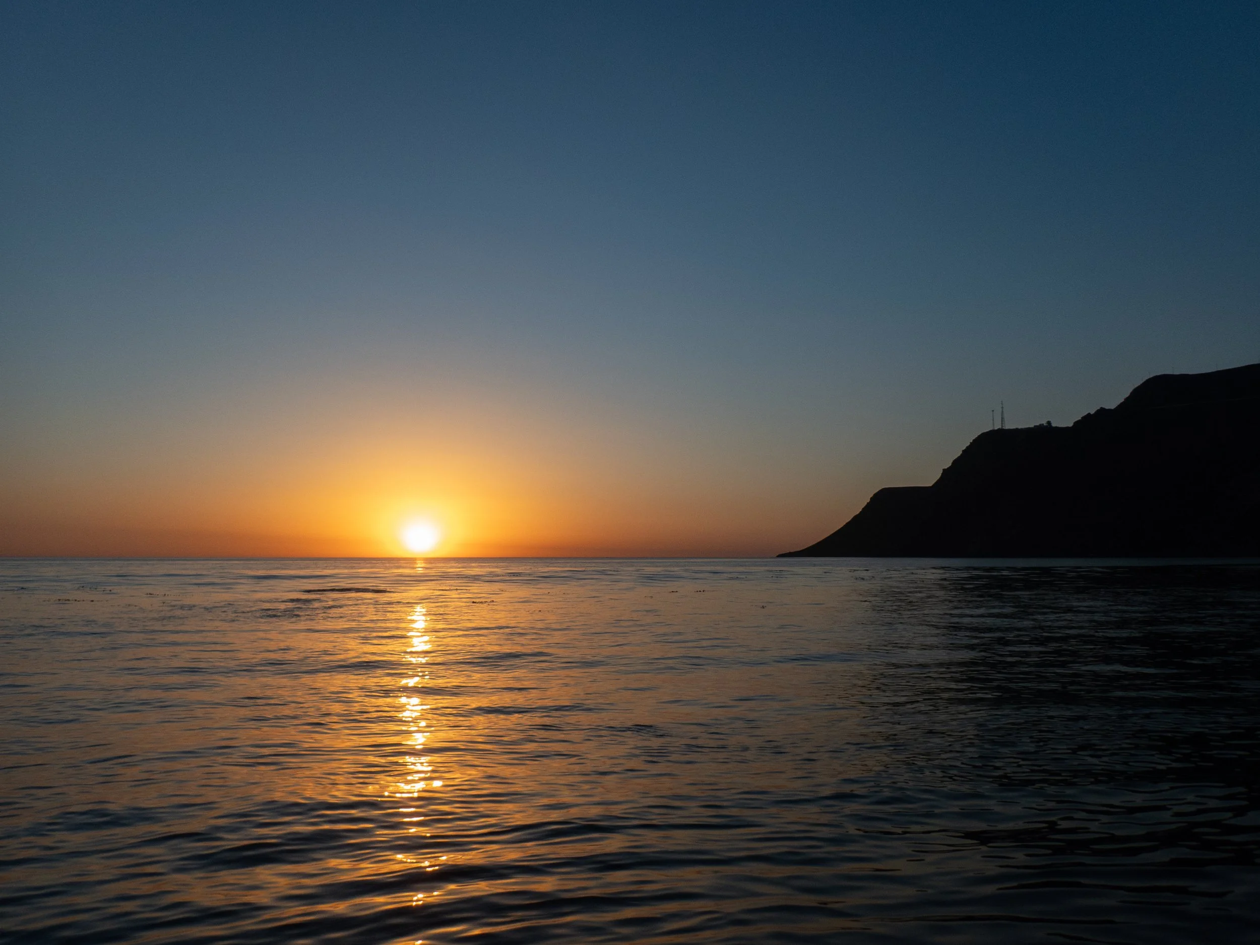 Sunset over the ocean with a silhouette of a hillside on the right side and calm water reflecting the sunset.
