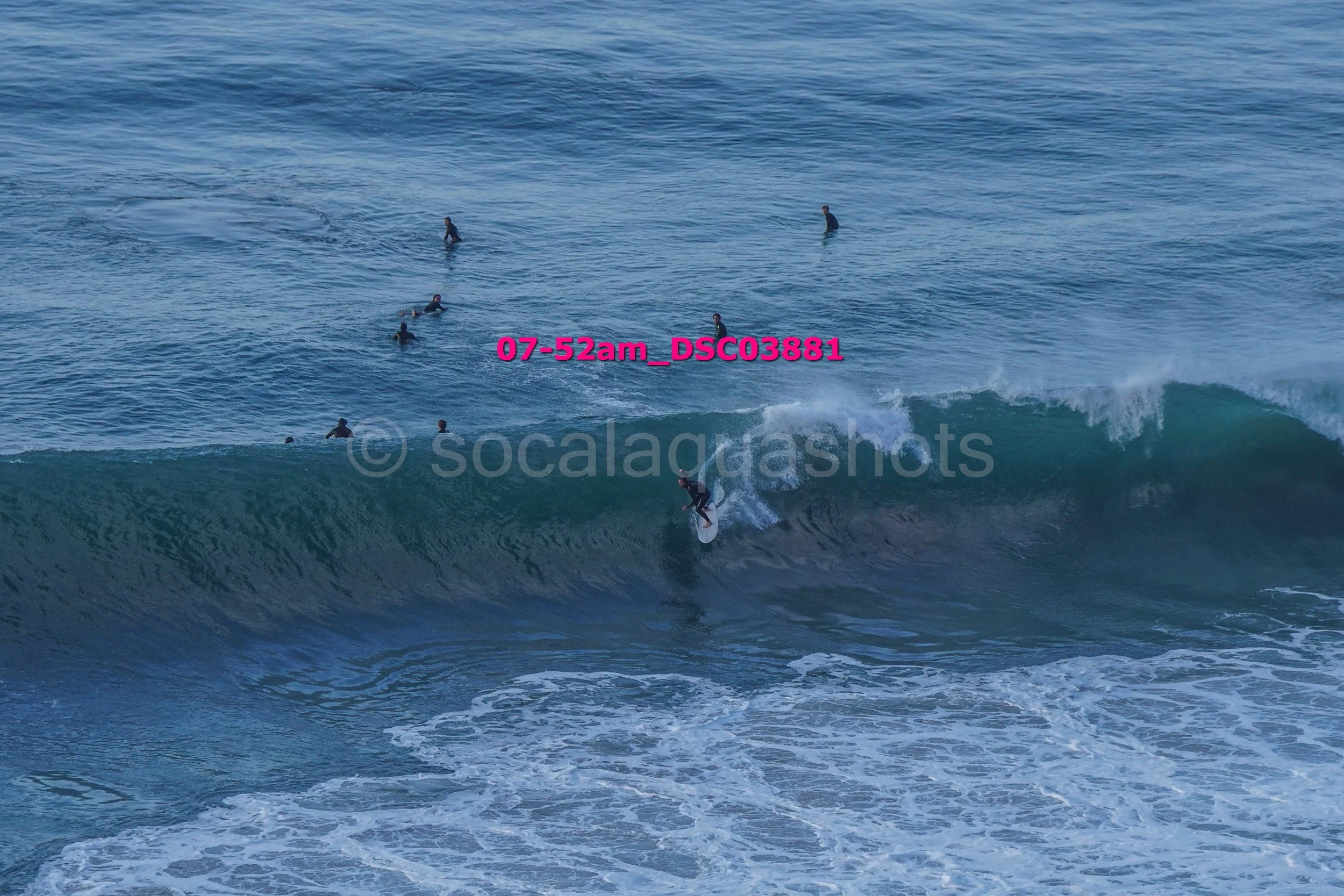 Surfer riding a wave with multiple people swimming and surfing in the background at the beach.
