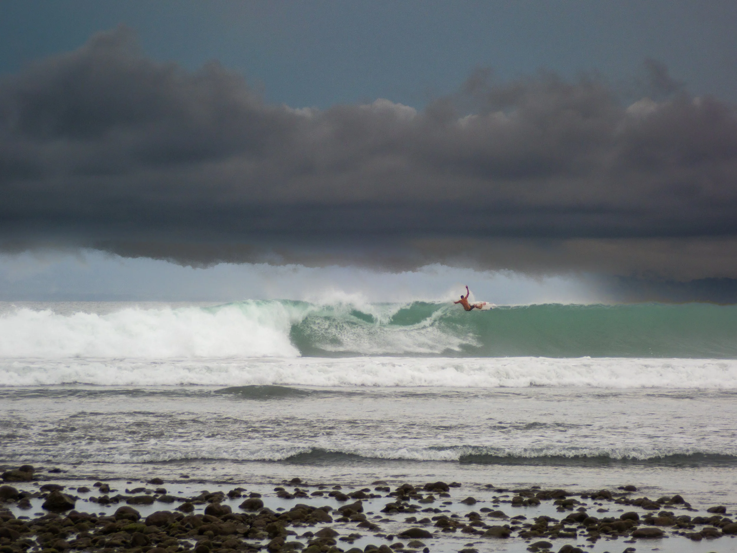 Surfer riding a wave under a cloudy sky near a rocky shoreline