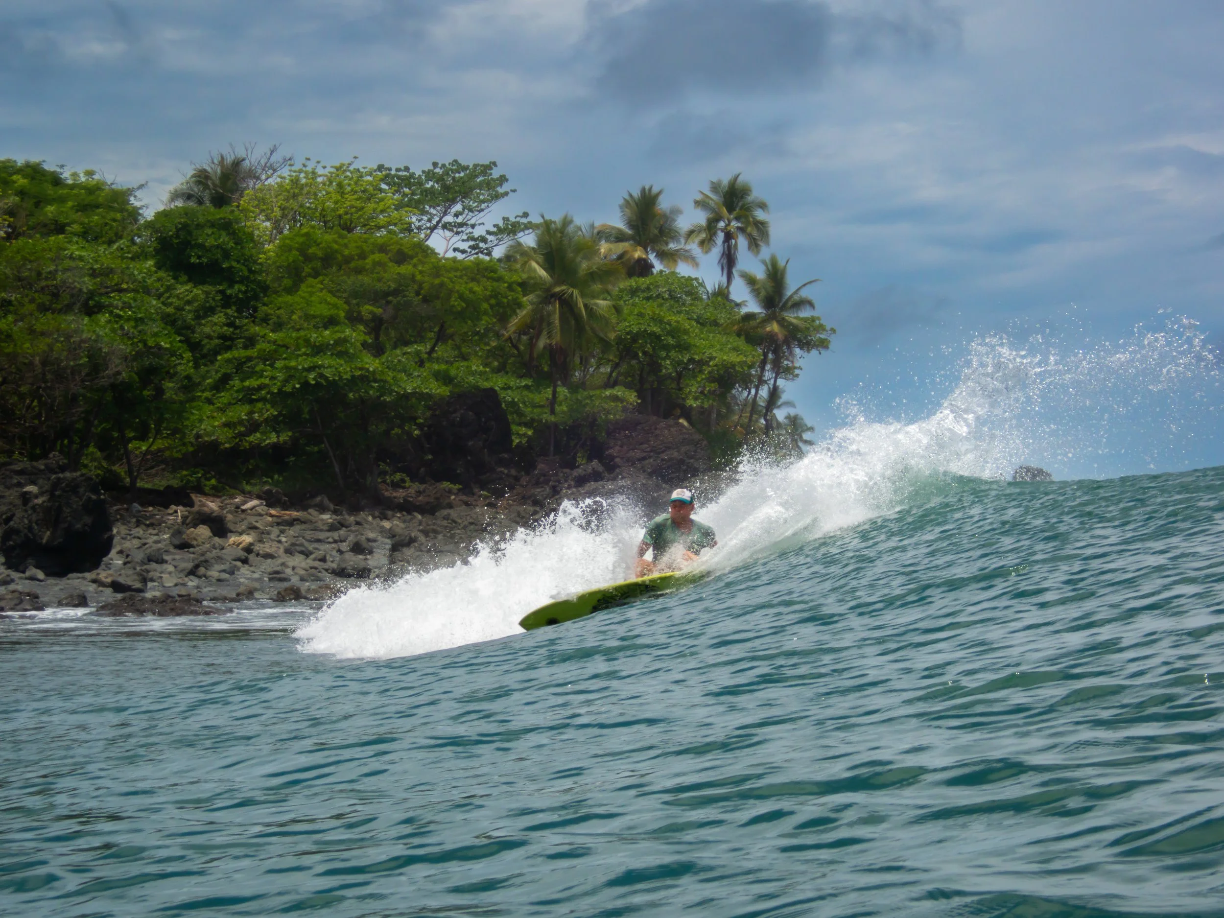Surfer riding a wave near a rocky shoreline with lush greenery.