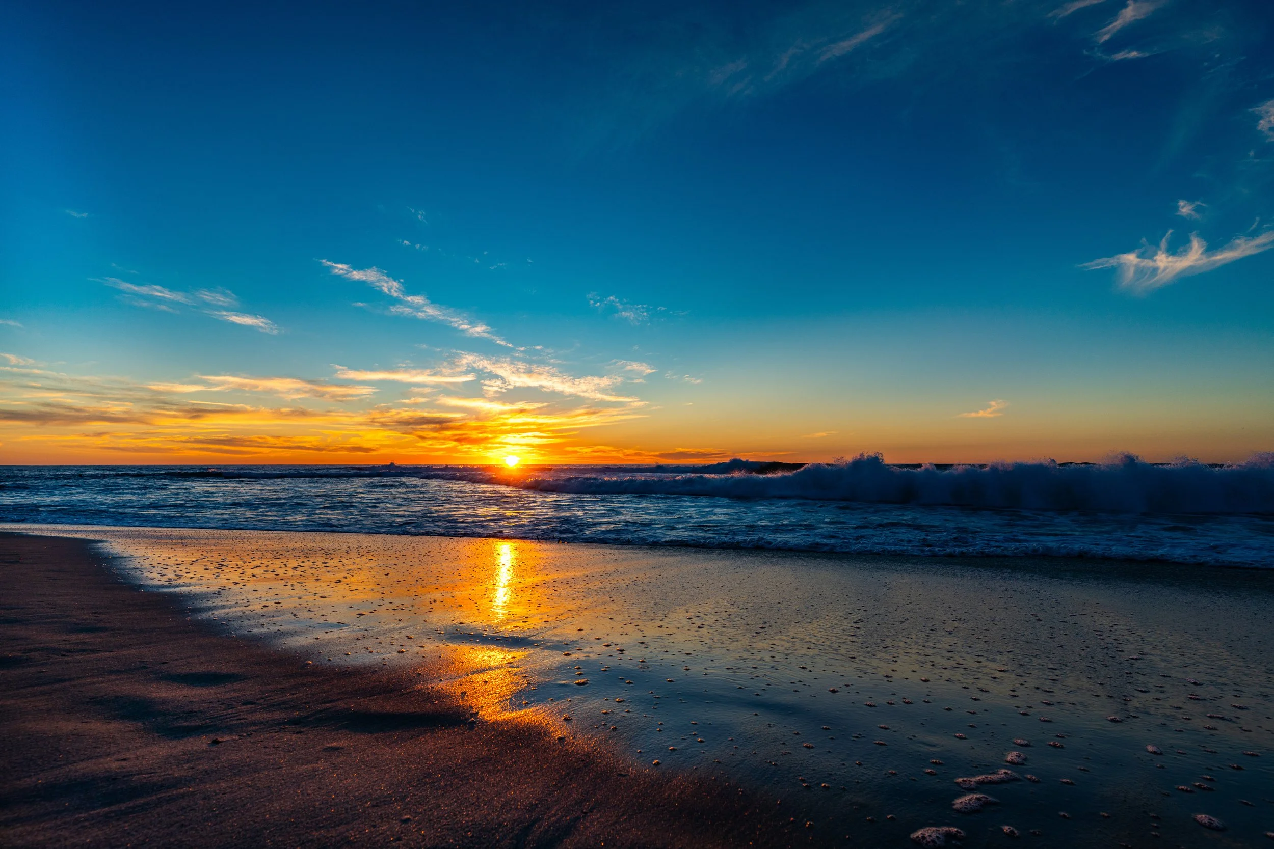 Sunset over the ocean with waves crashing onto the sandy beach, colorful sky with clouds.