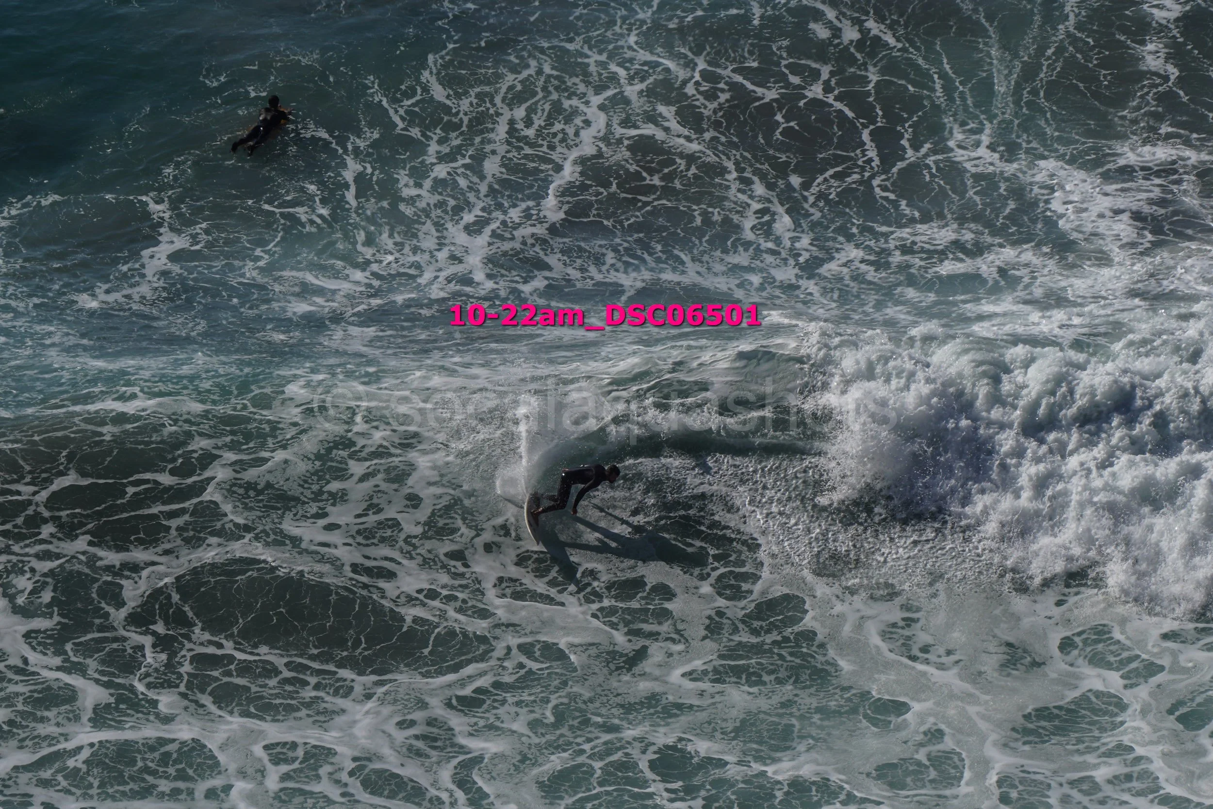 A person surfing on a wave in the ocean with another surfer visible further away. The image has a pink timestamp overlay reading '10-22am_DSC6501'.