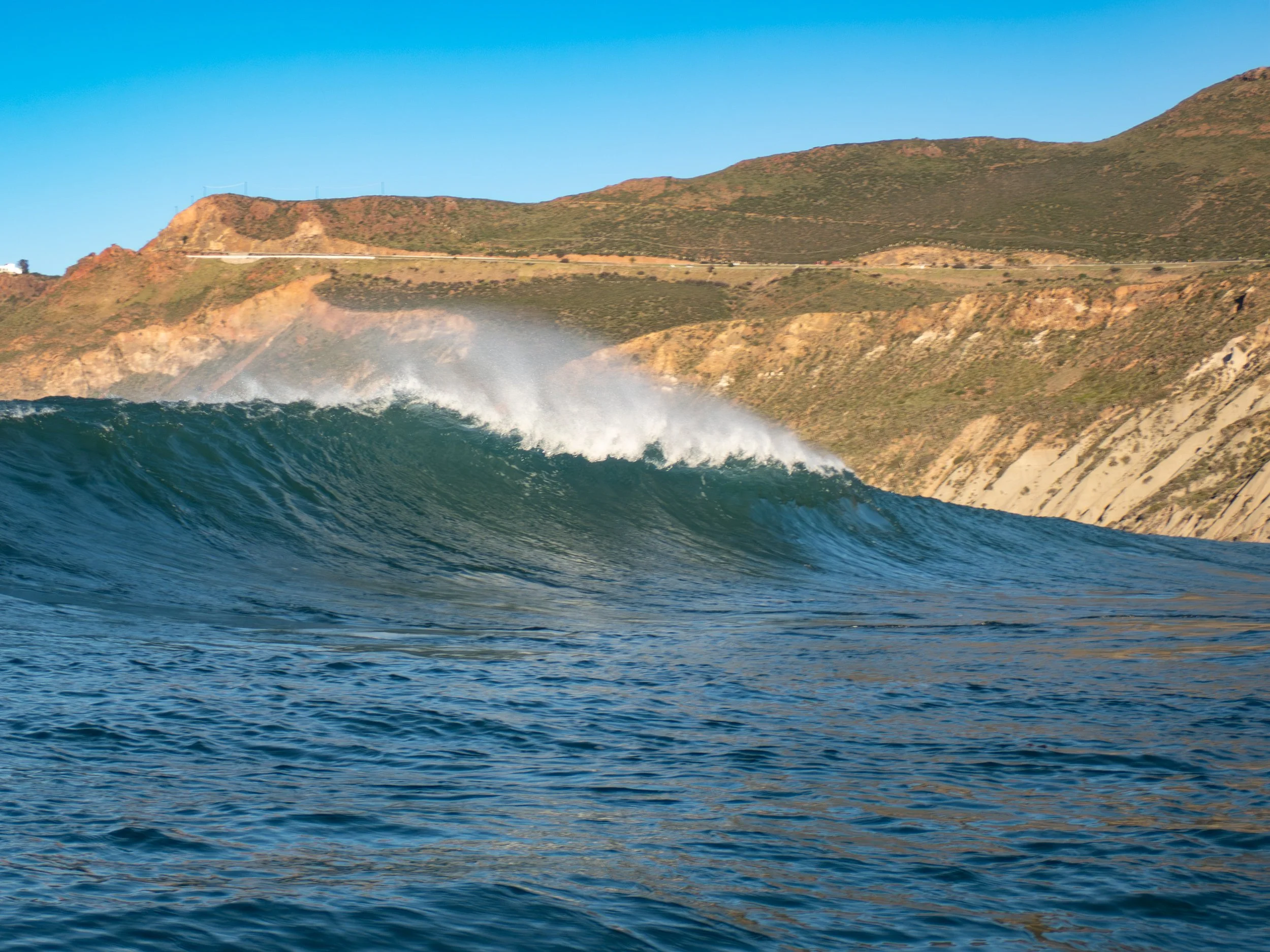 Ocean wave approaching shoreline with hills in the background under clear blue sky.