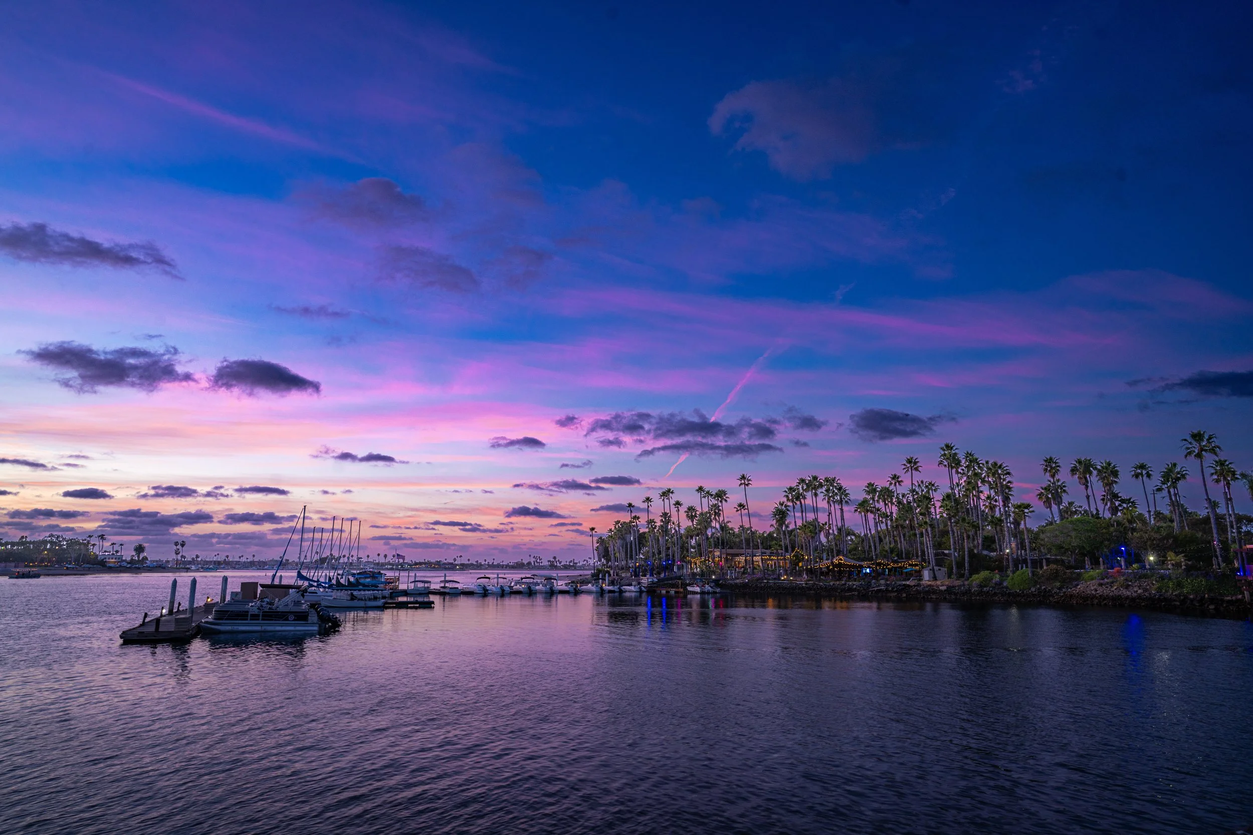A harbor at sunset with boats docked, tall palm trees along the shoreline, and a colorful sky with pink and purple clouds.