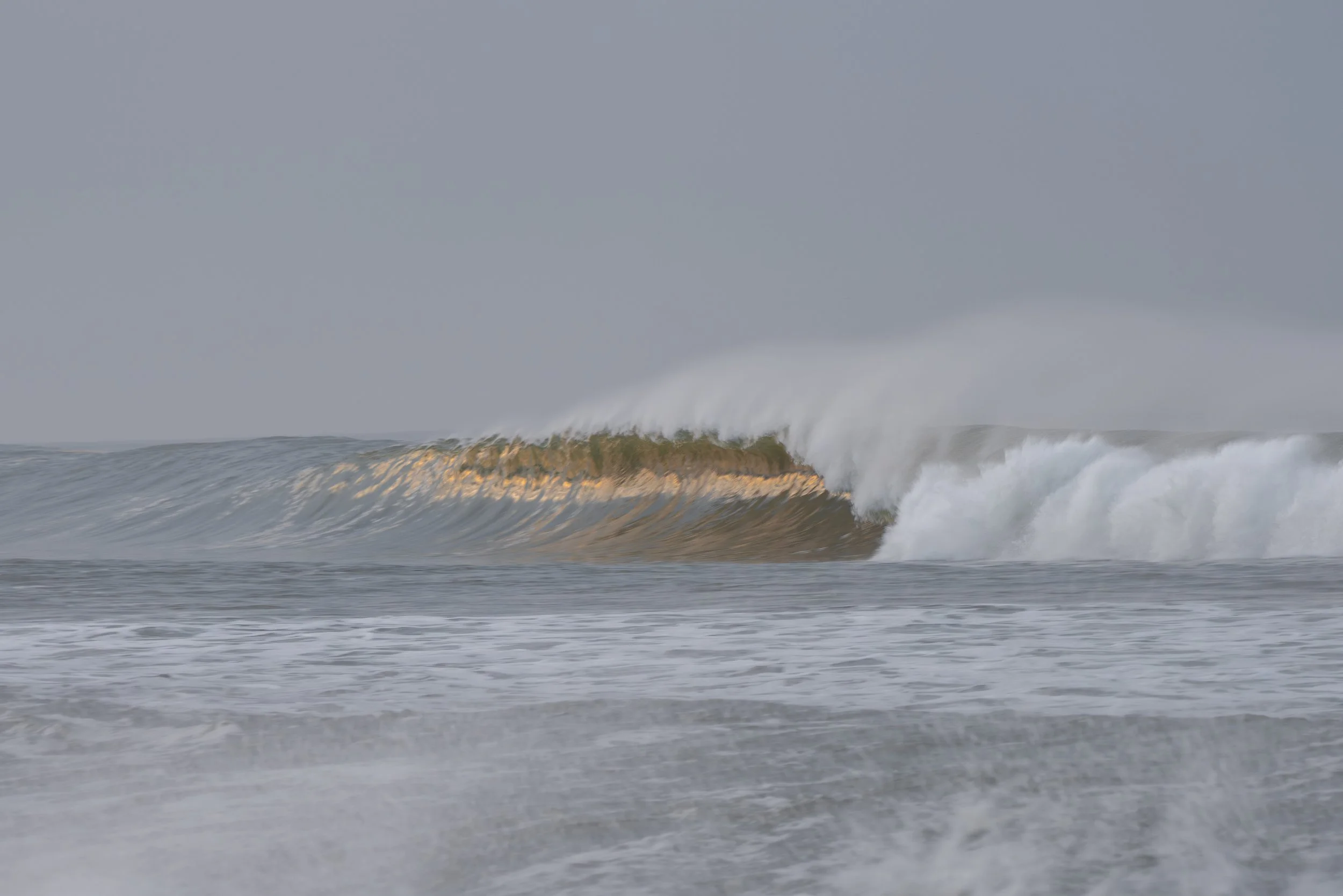 Ocean waves crashing, with a large wave breaking and spray in the air against a cloudy sky.