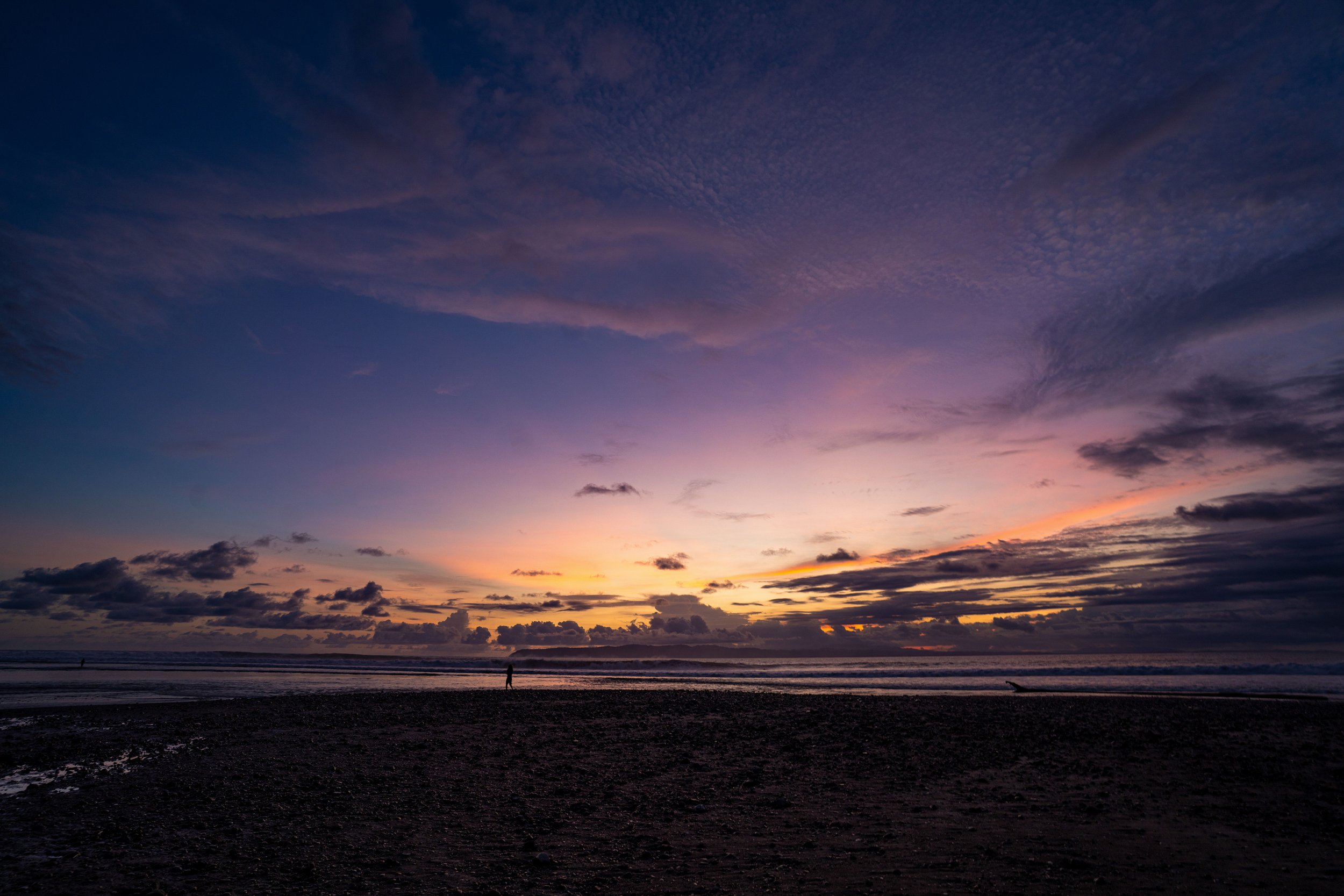 A sunset at the beach with a person walking along the shore and a colorful sky with clouds.