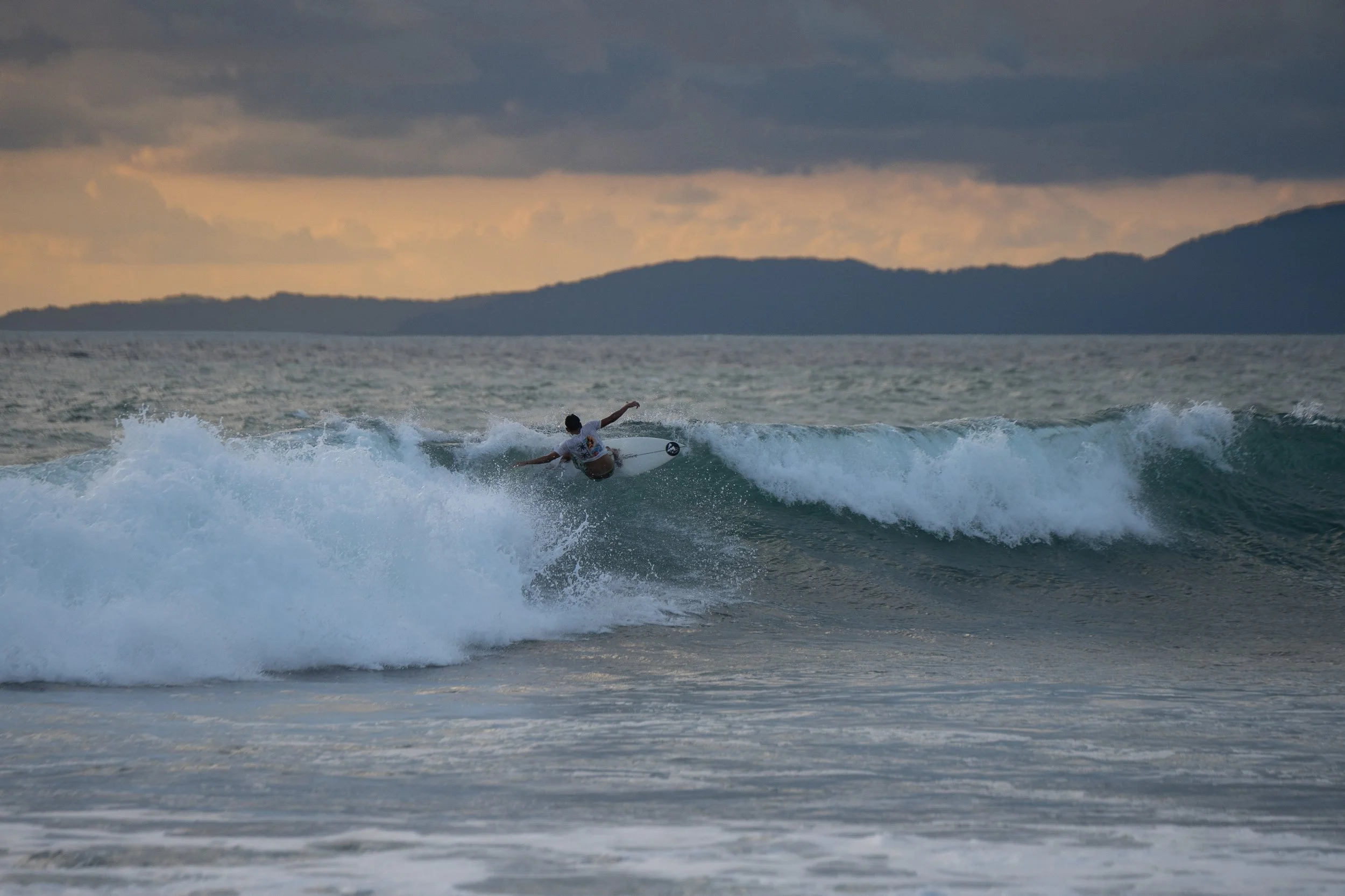 A person surfing on a wave in the ocean during sunset, with a distant mountainous landmass in the background.