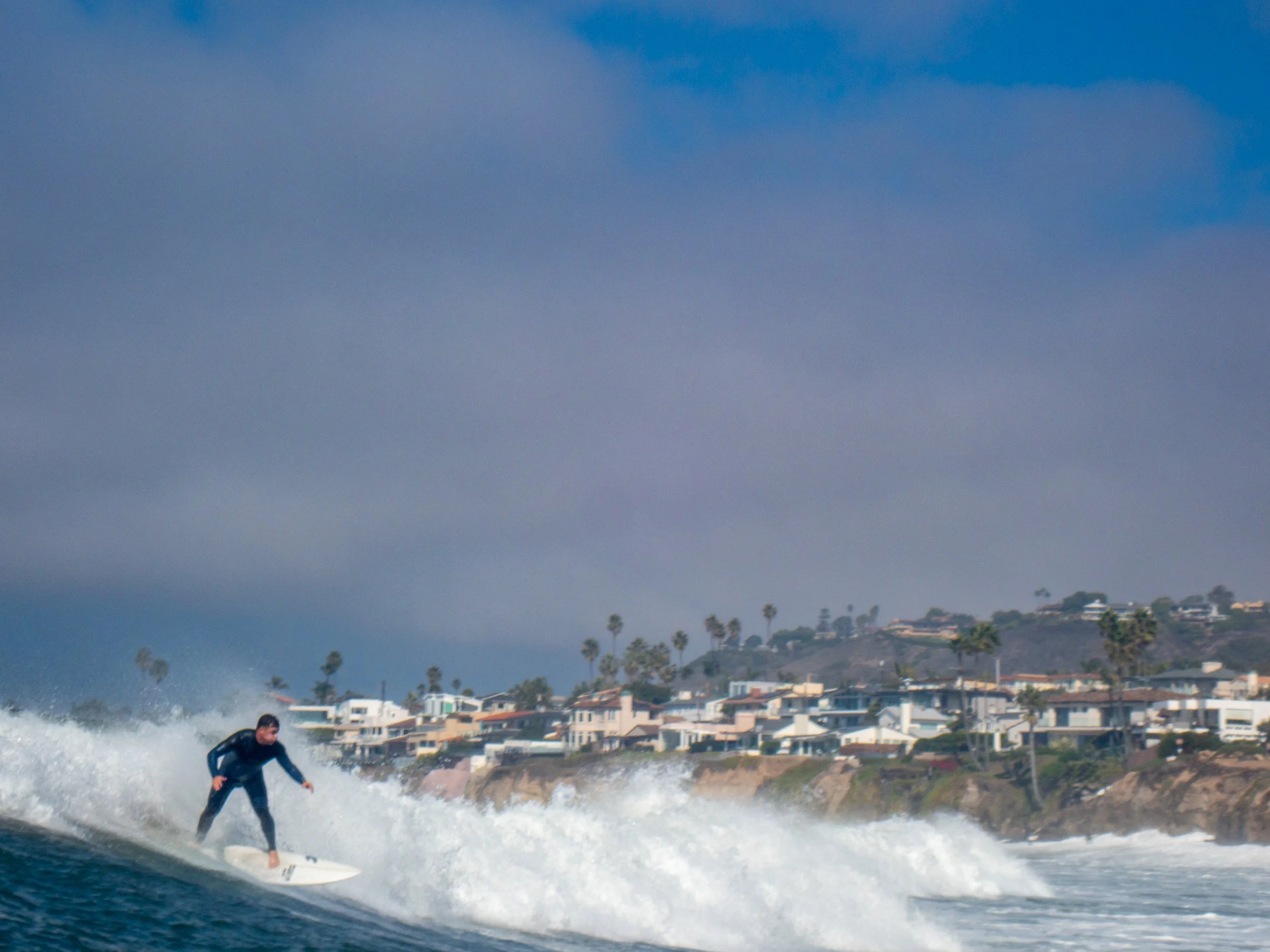 Person surfing on a wave near the shoreline with houses and palm trees in the background under a cloudy sky.