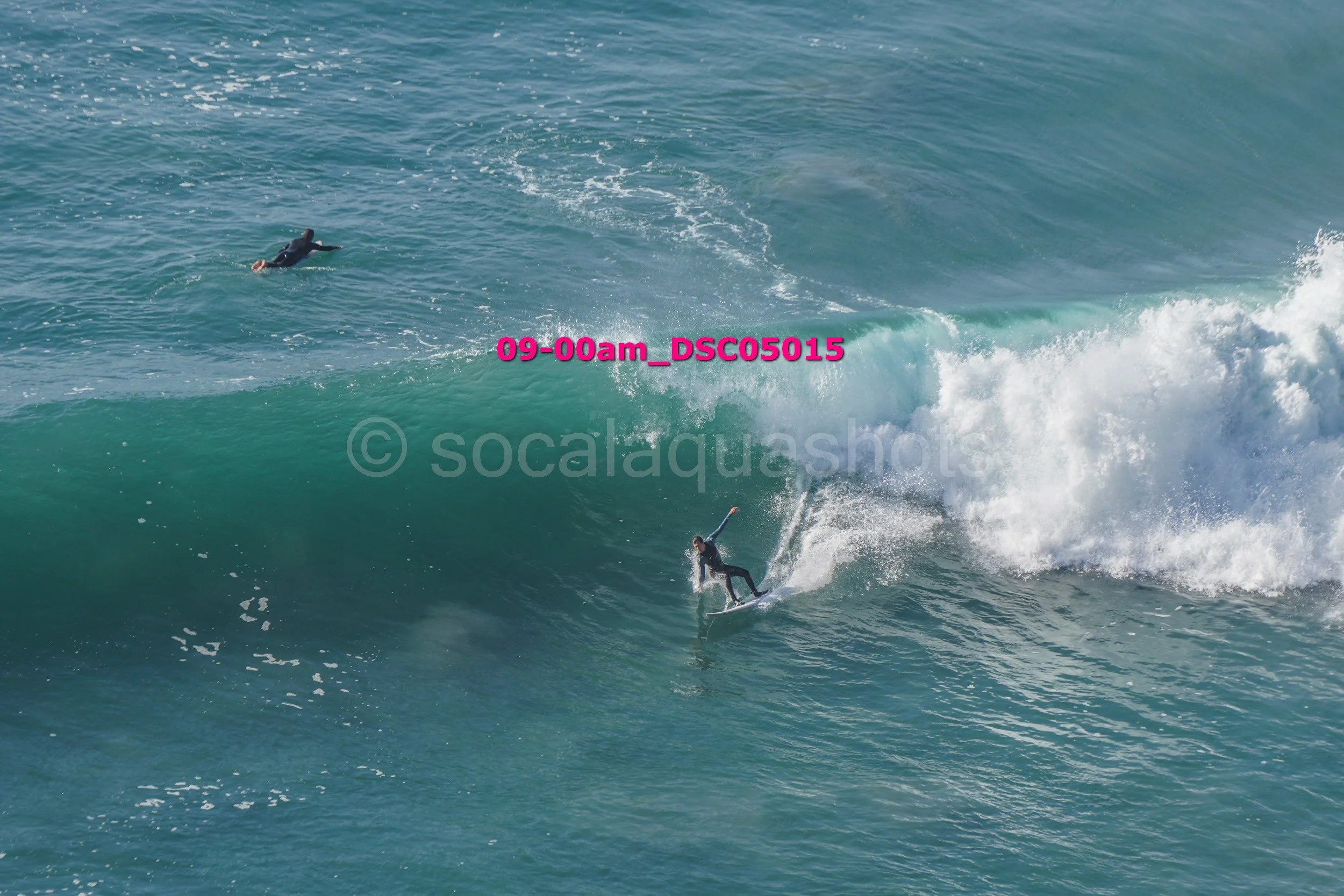 A surfer riding a wave with another person swimming in the background. The water is blue and white with visible spray.
