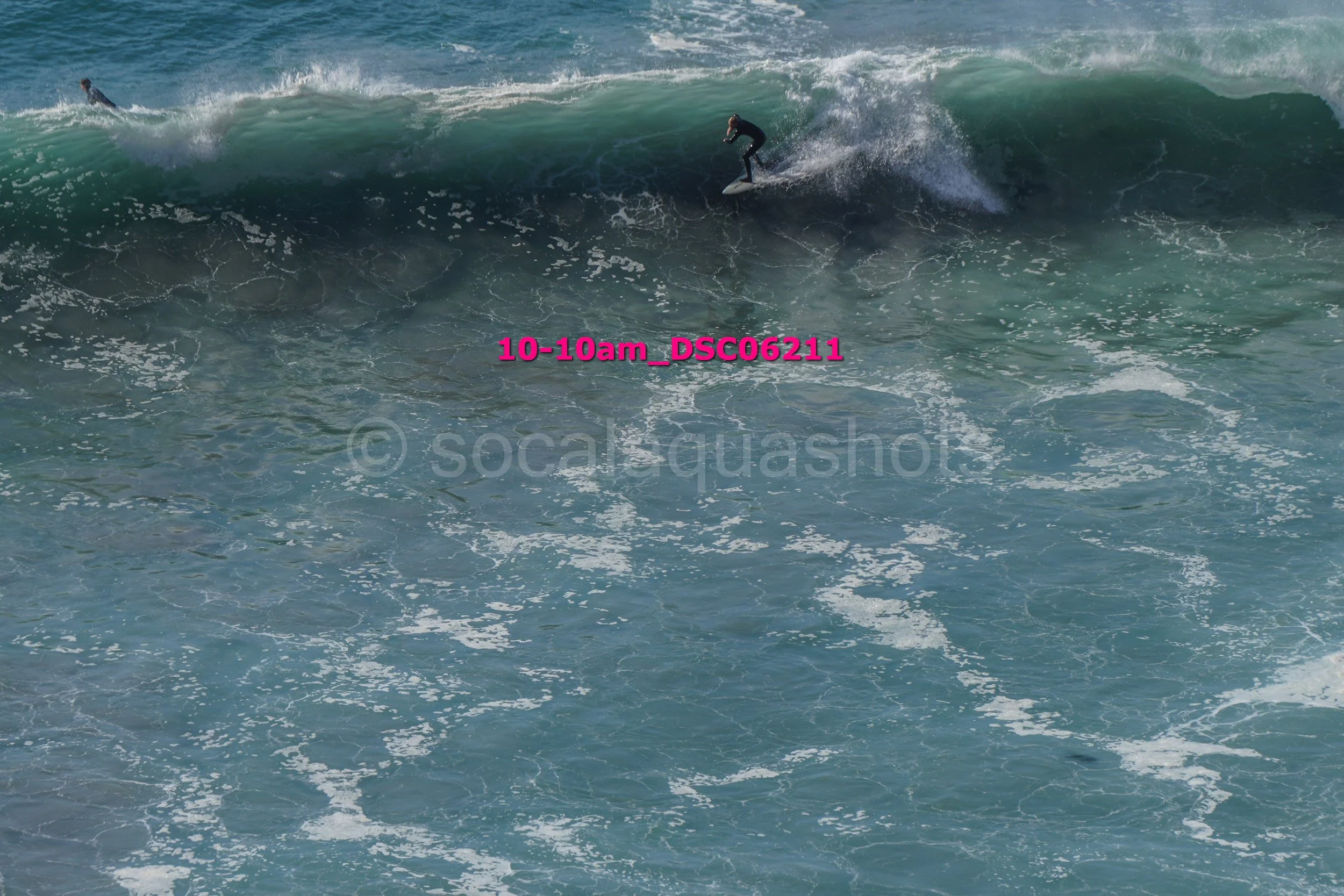 A person surfing on a large wave in the ocean, with another surfer visible in the background.