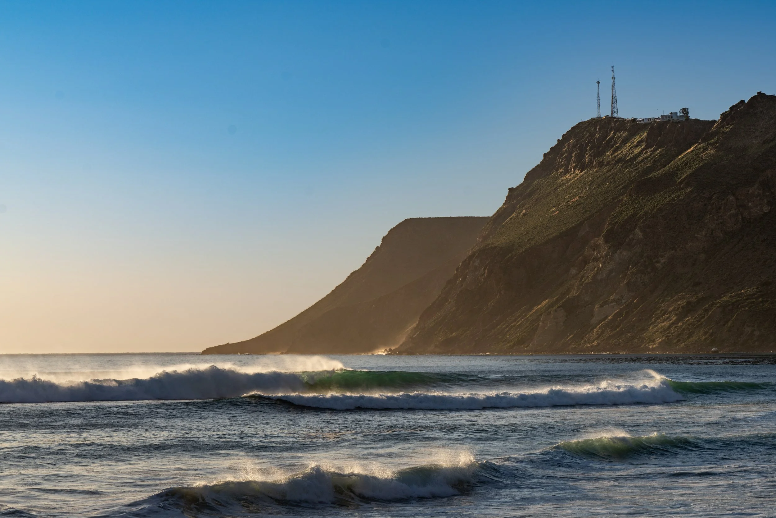 Ocean waves breaking near a rocky shoreline with a steep hillside and communication towers on top under a clear blue sky at sunset.