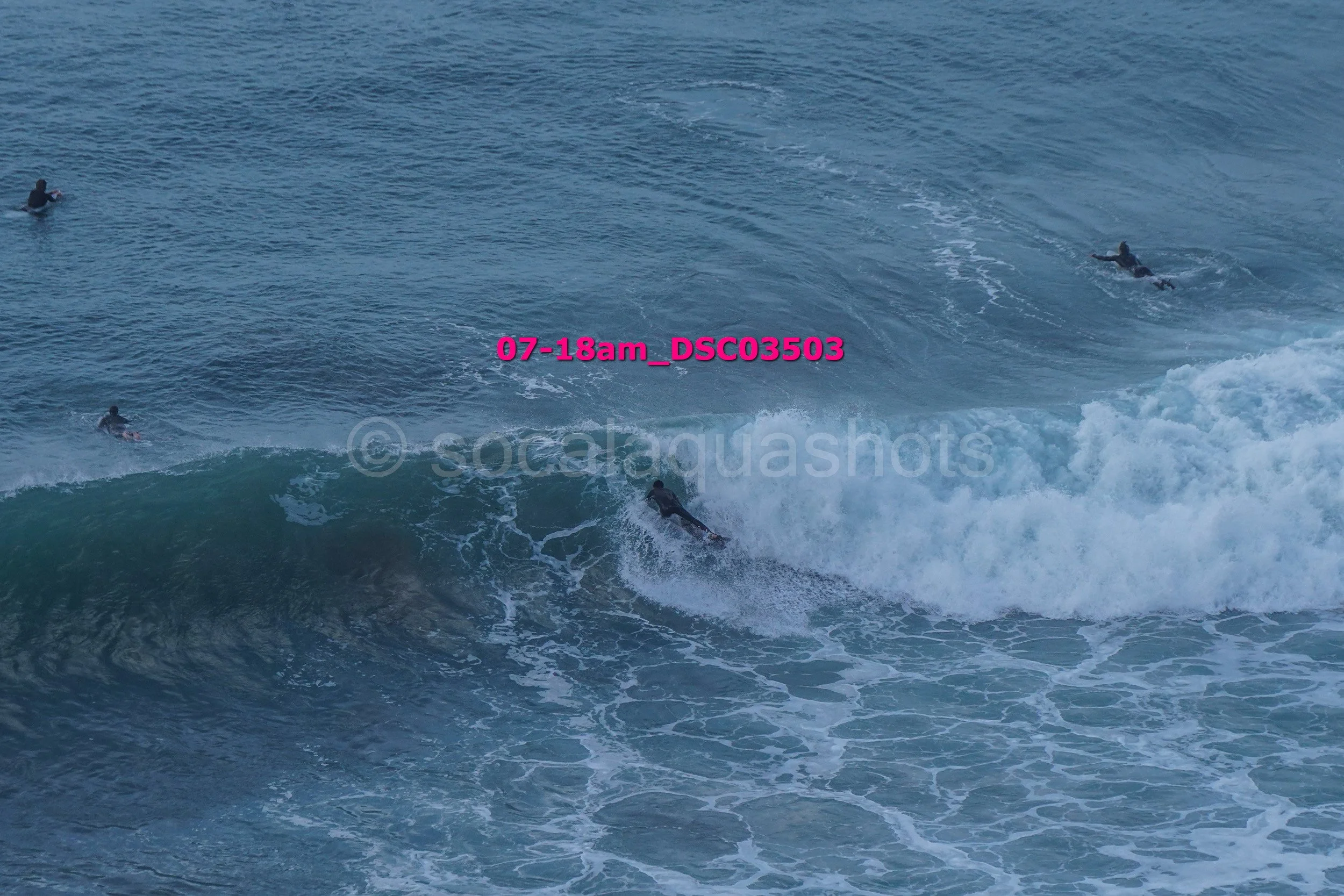 Surfers riding a wave in the ocean with three surfers visible, one paddling and others surfing.