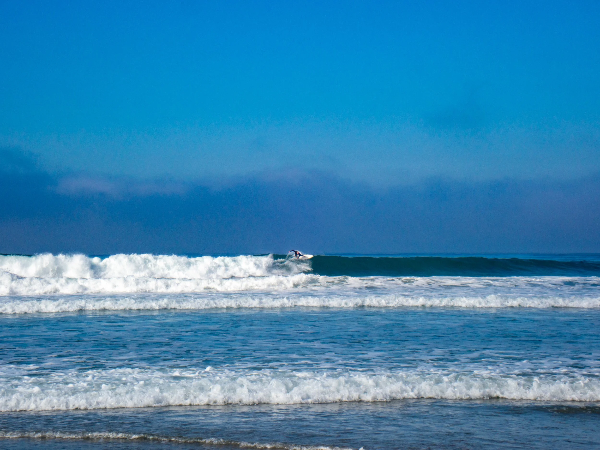 A surfer riding a wave in the ocean under a partly cloudy blue sky.
