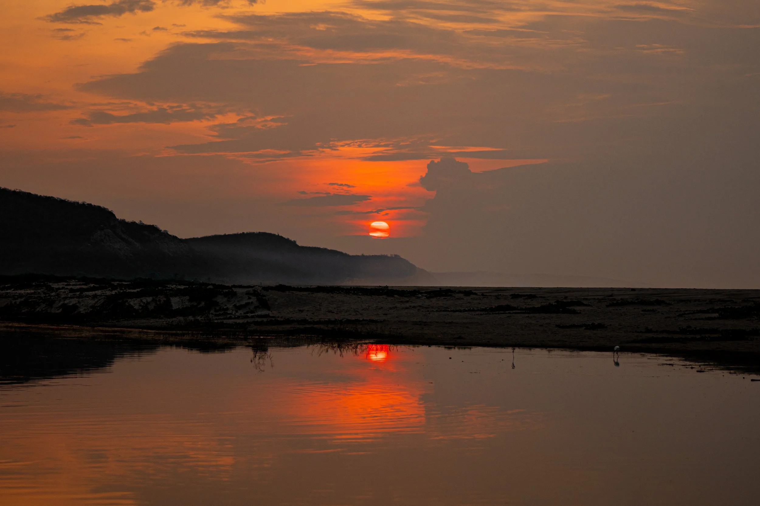 A sunset over a body of water with mountains in the distance, with the sky filled with orange and pink clouds, and the sun partially visible near the horizon.