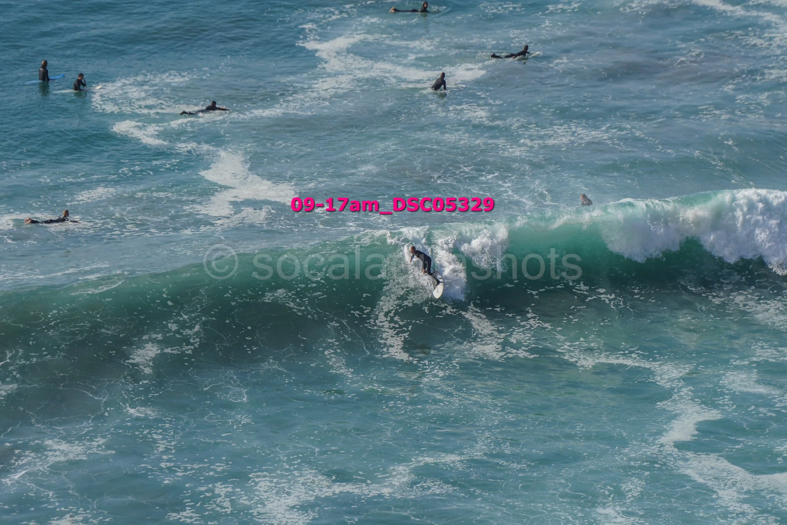 Surfer riding a wave in the ocean surrounded by several people swimming and surfing.