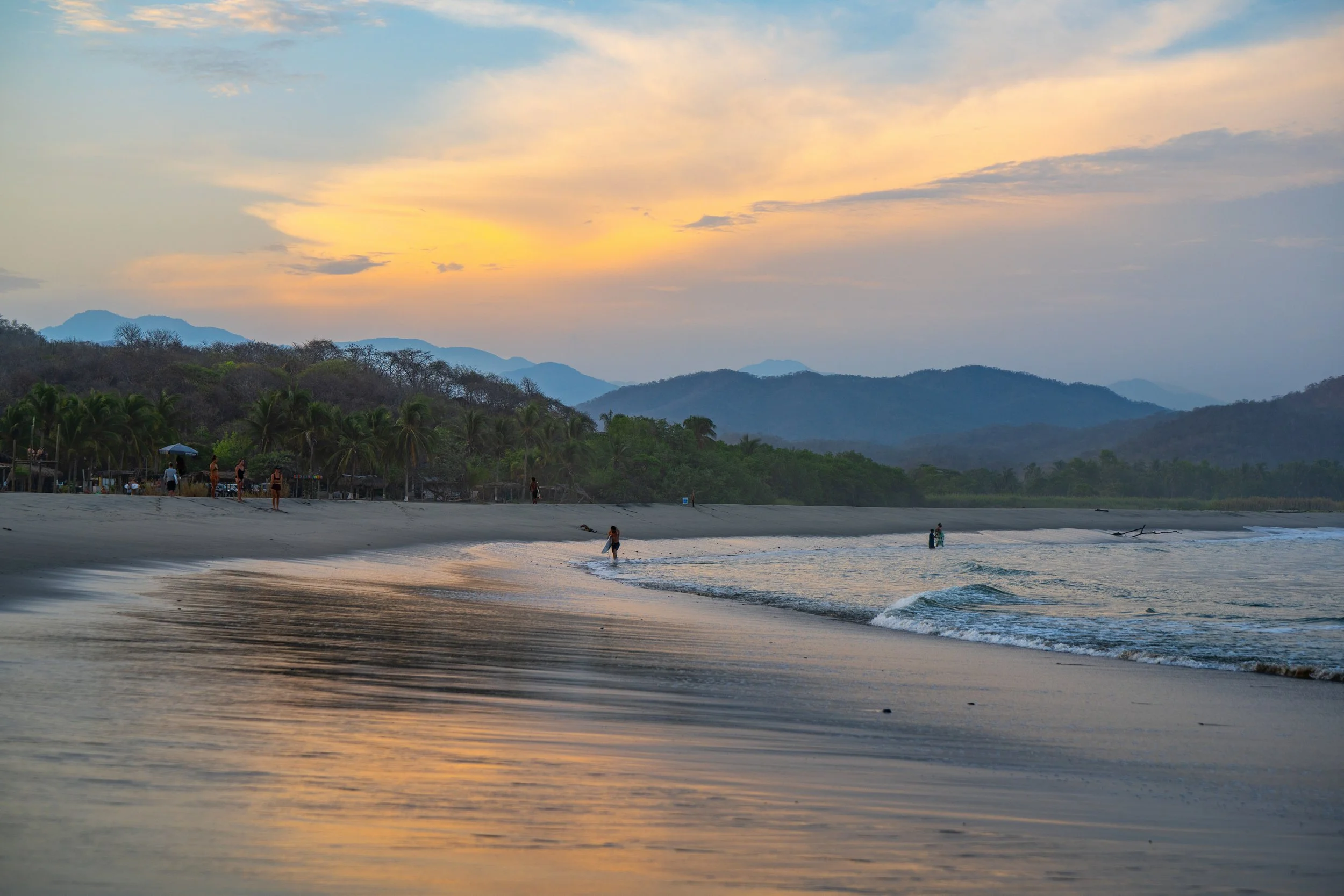 Sunset over a beach with gentle waves, a few people walking along the shore, lush green hills and mountains in the background, and a partly cloudy sky with soft yellow, orange, and blue hues.