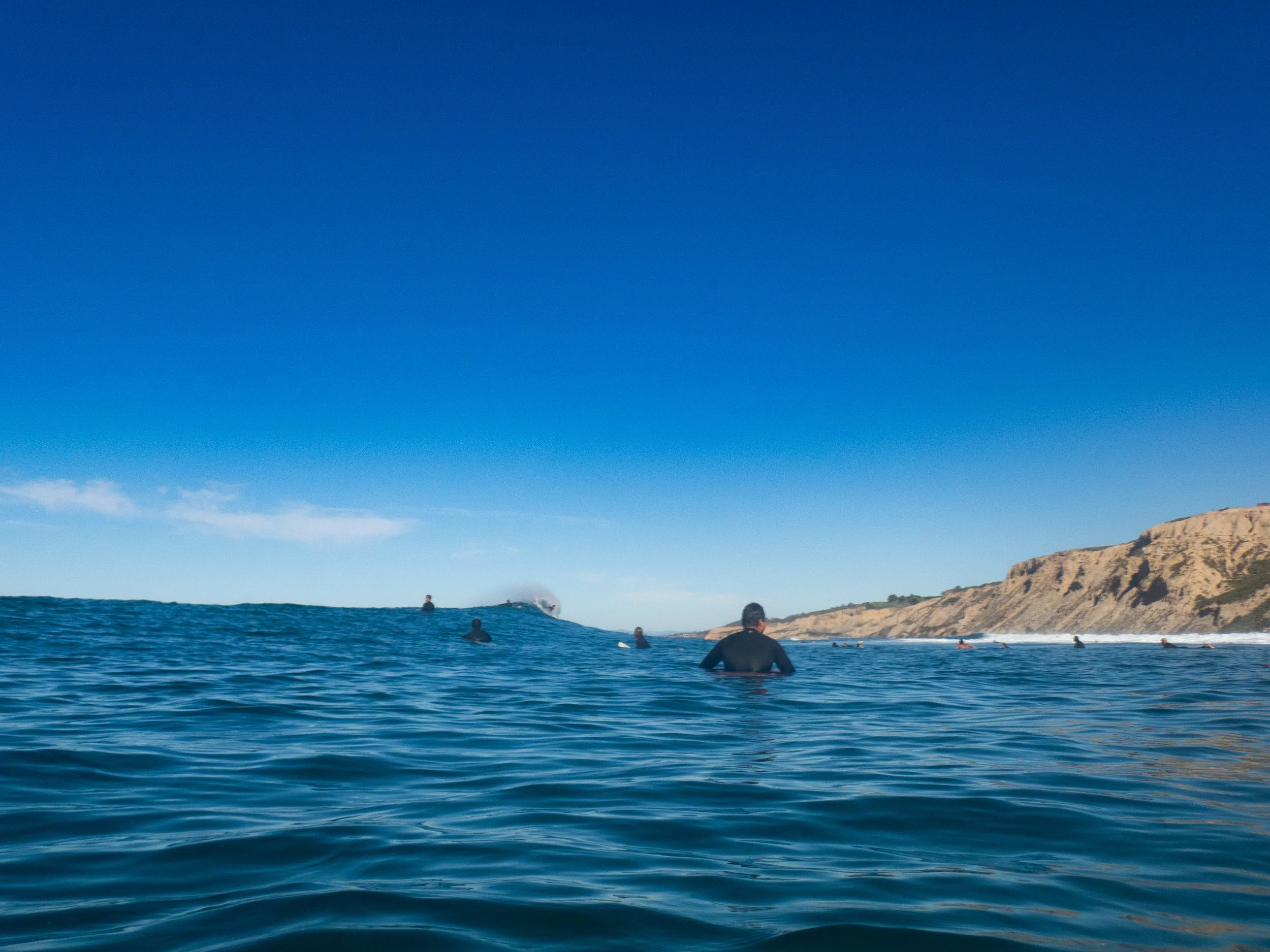 People surfing and swimming in the ocean with cliffs in the background under a clear blue sky.