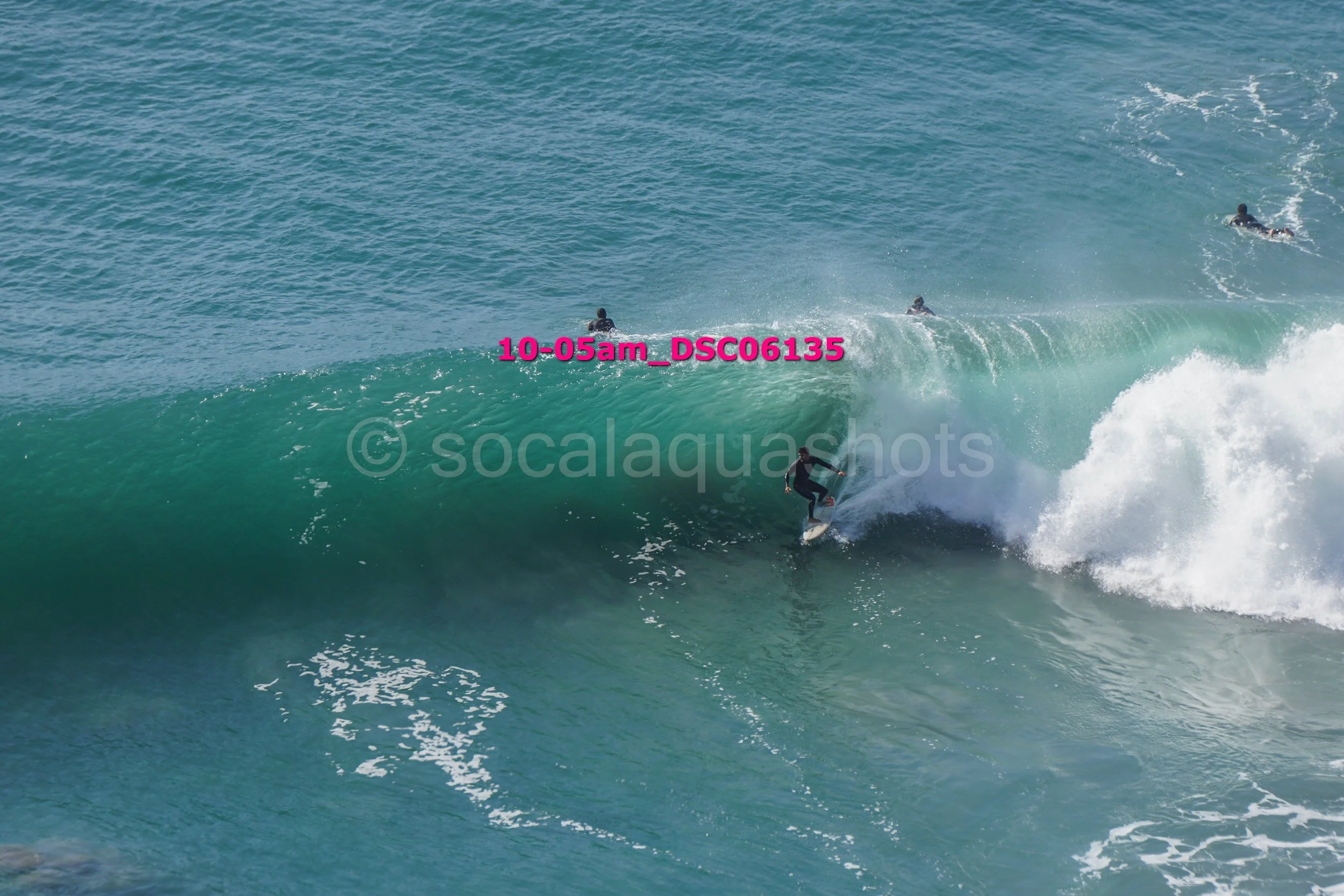 A surfer riding a large wave, with other surfers in the water nearby, in an ocean setting.
