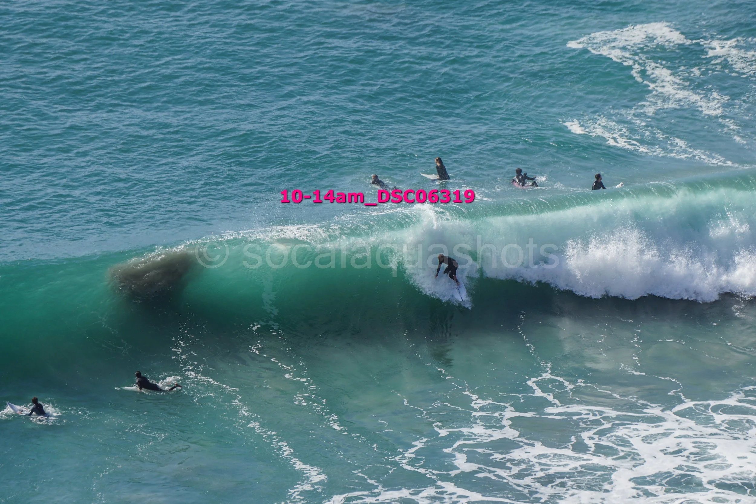 Surfer riding a large ocean wave with several other surfers in the water nearby.