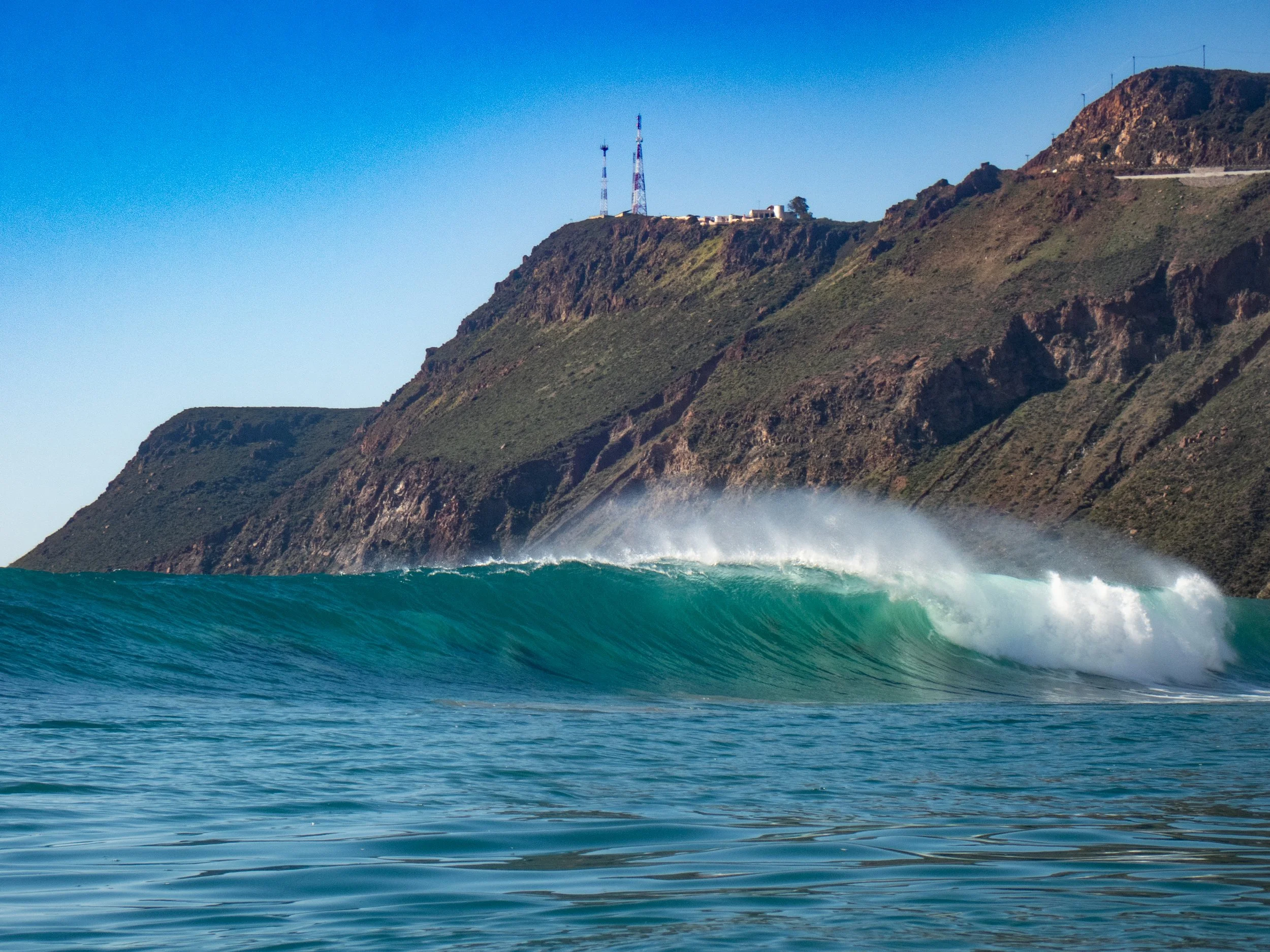A large ocean wave rolling towards the shore with greenish-blue water, with a rugged hillside in the background featuring antennas on top of the hill.