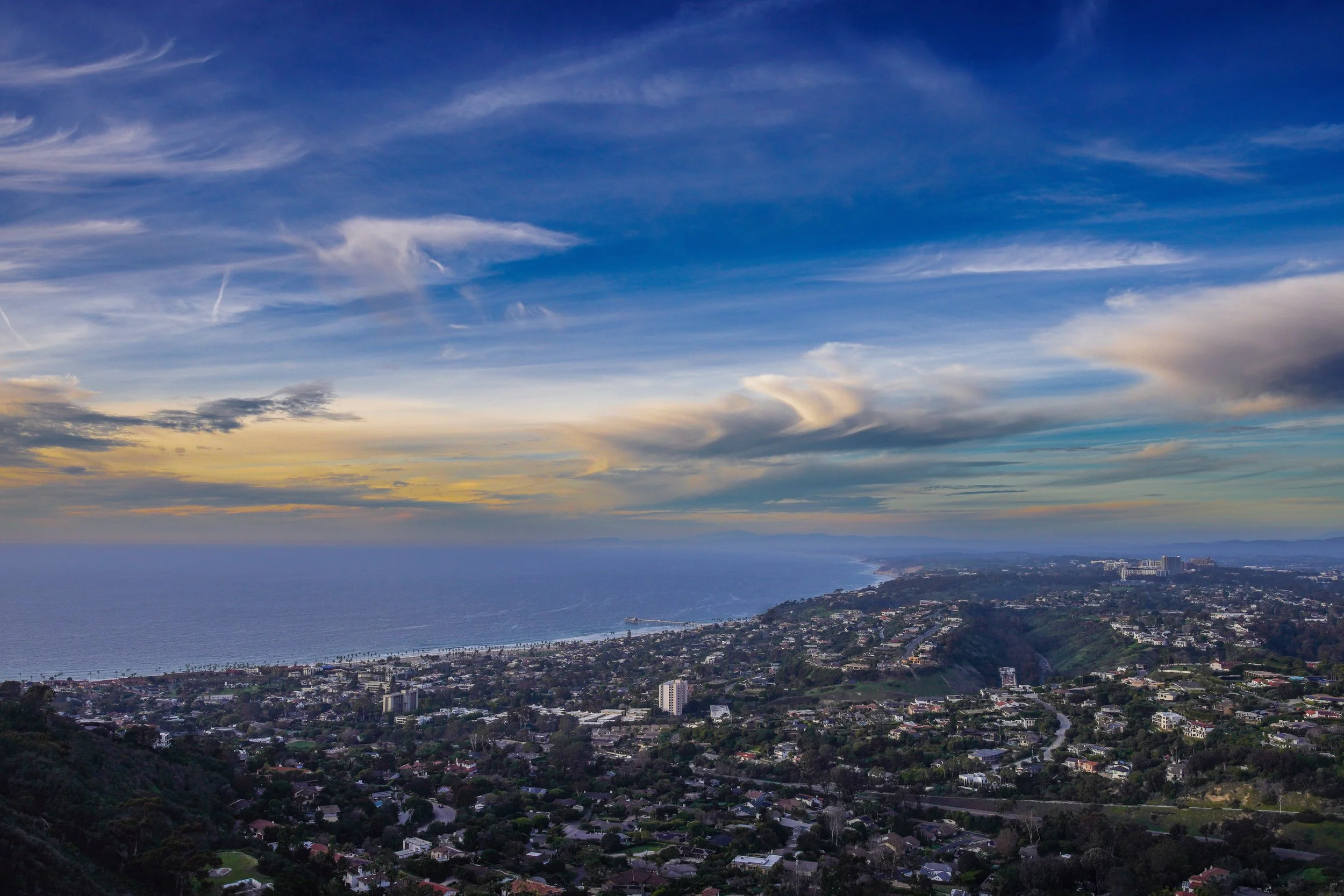 A panoramic view of a coastal city with houses, buildings, and hills, overlooking the ocean under a partly cloudy sky.
