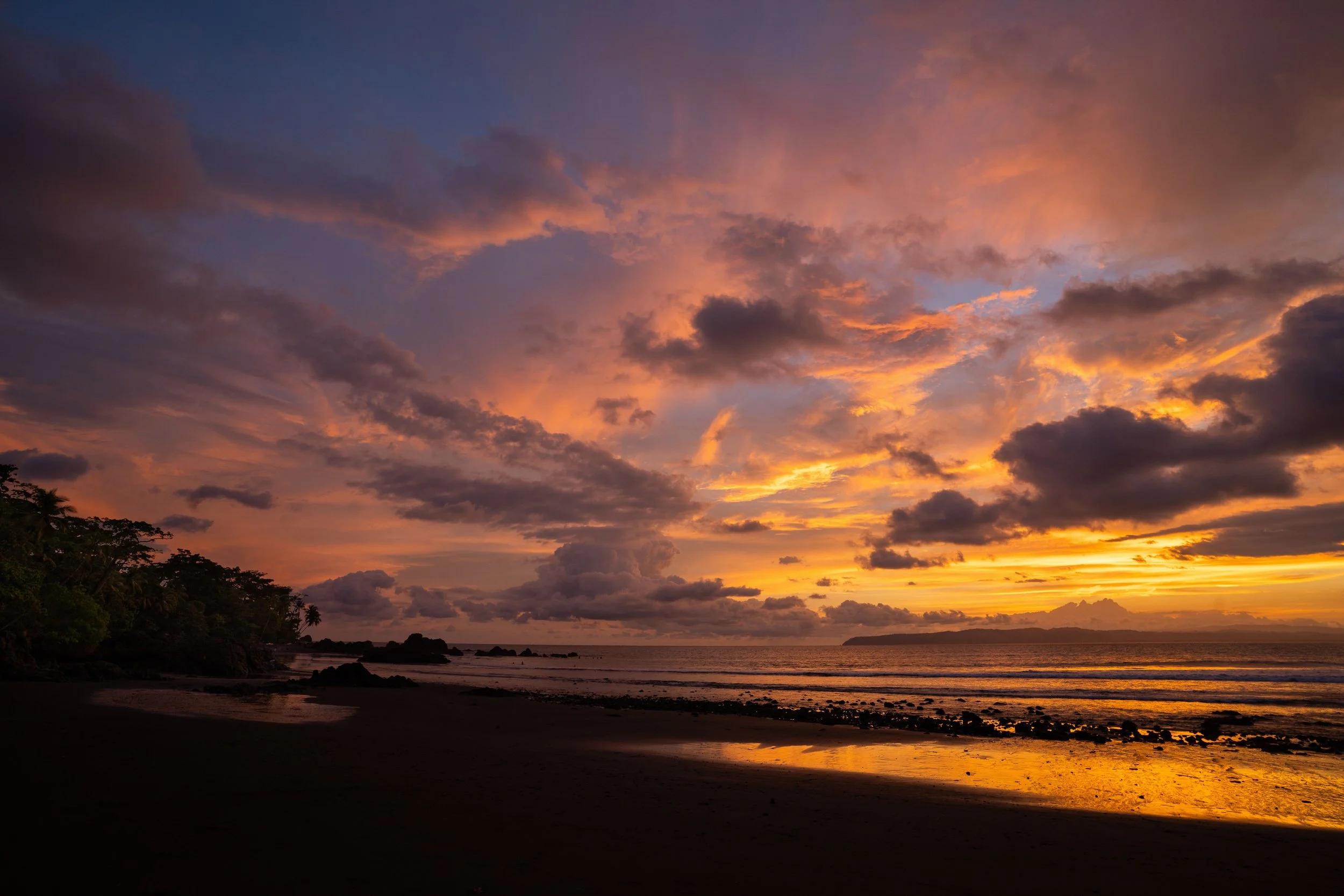 Sunset over a beach with a colorful sky filled with clouds, silhouetted trees, and the calm ocean reflecting the sky's colors.