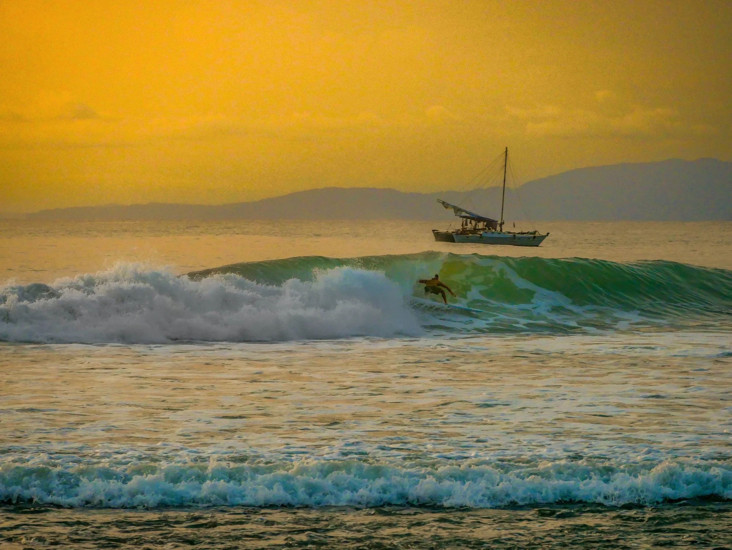 Surfer riding a wave with a sailboat in the background during sunset