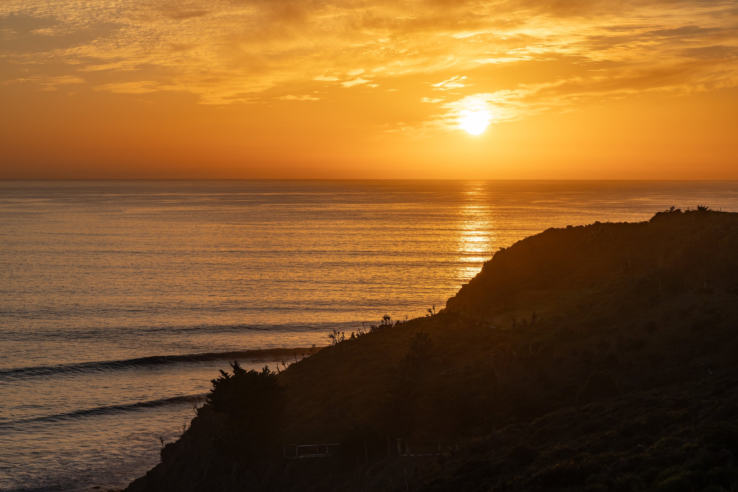 Sunset over the ocean with a rocky headland in the foreground, the sky filled with golden clouds and the sun reflecting on the water.