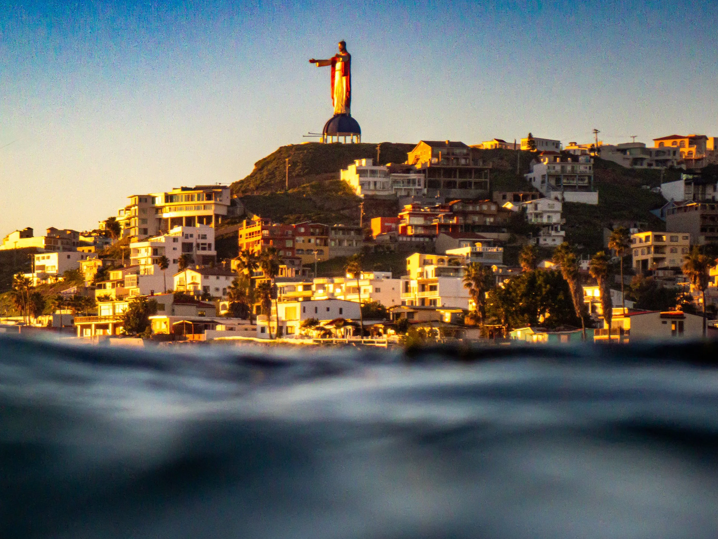 View of the cityscape with the Christ the Redeemer statue on a hillside during sunset, seen from over water.