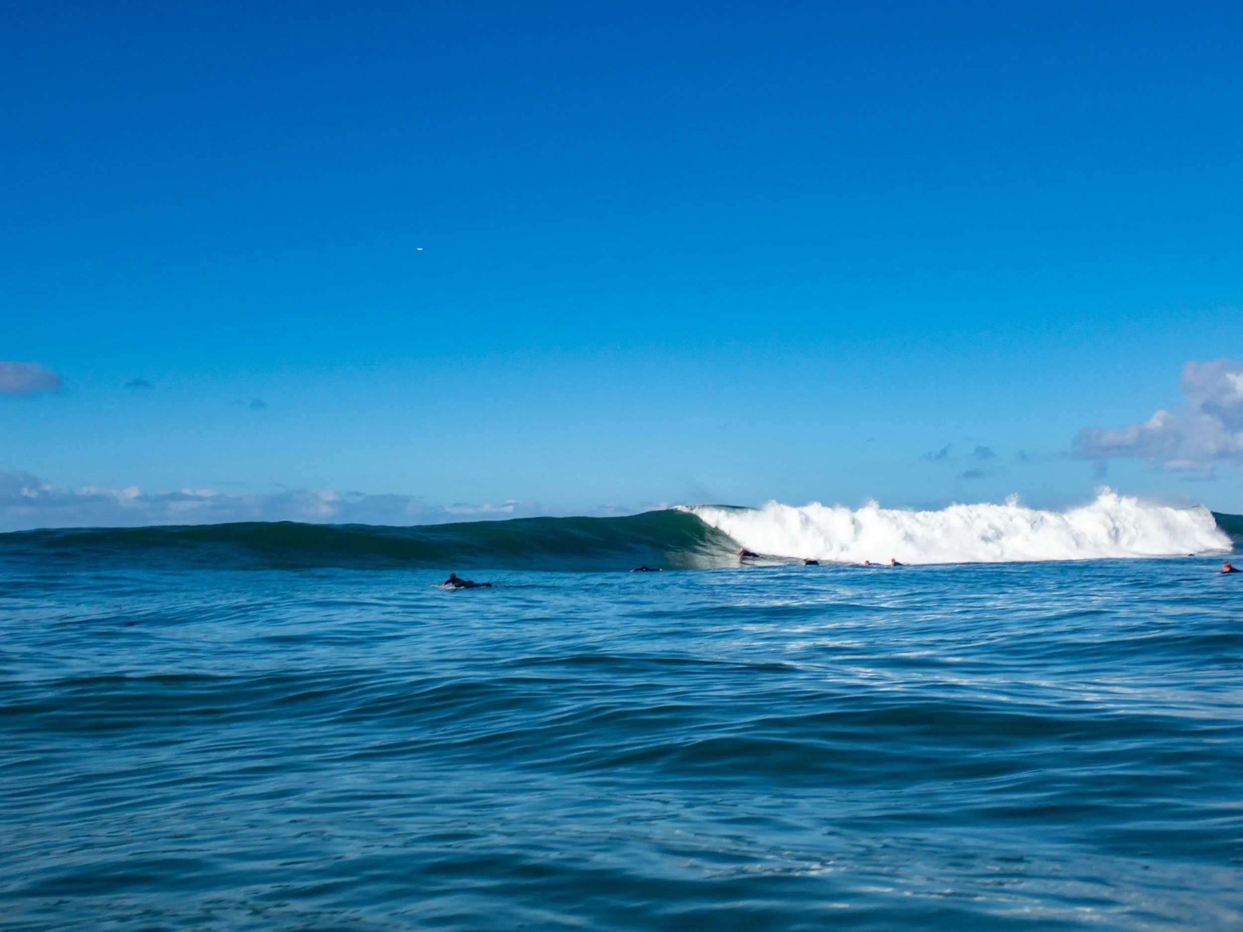 Surfers riding a large wave in the ocean under a clear blue sky.