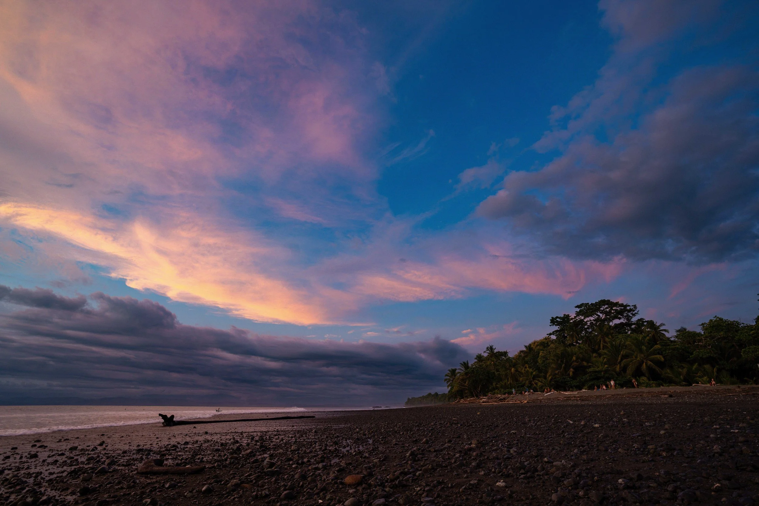 A beach at sunset with a colorful sky of pink, purple, and blue clouds, silhouetted trees, and a person sitting on a log near the shore.