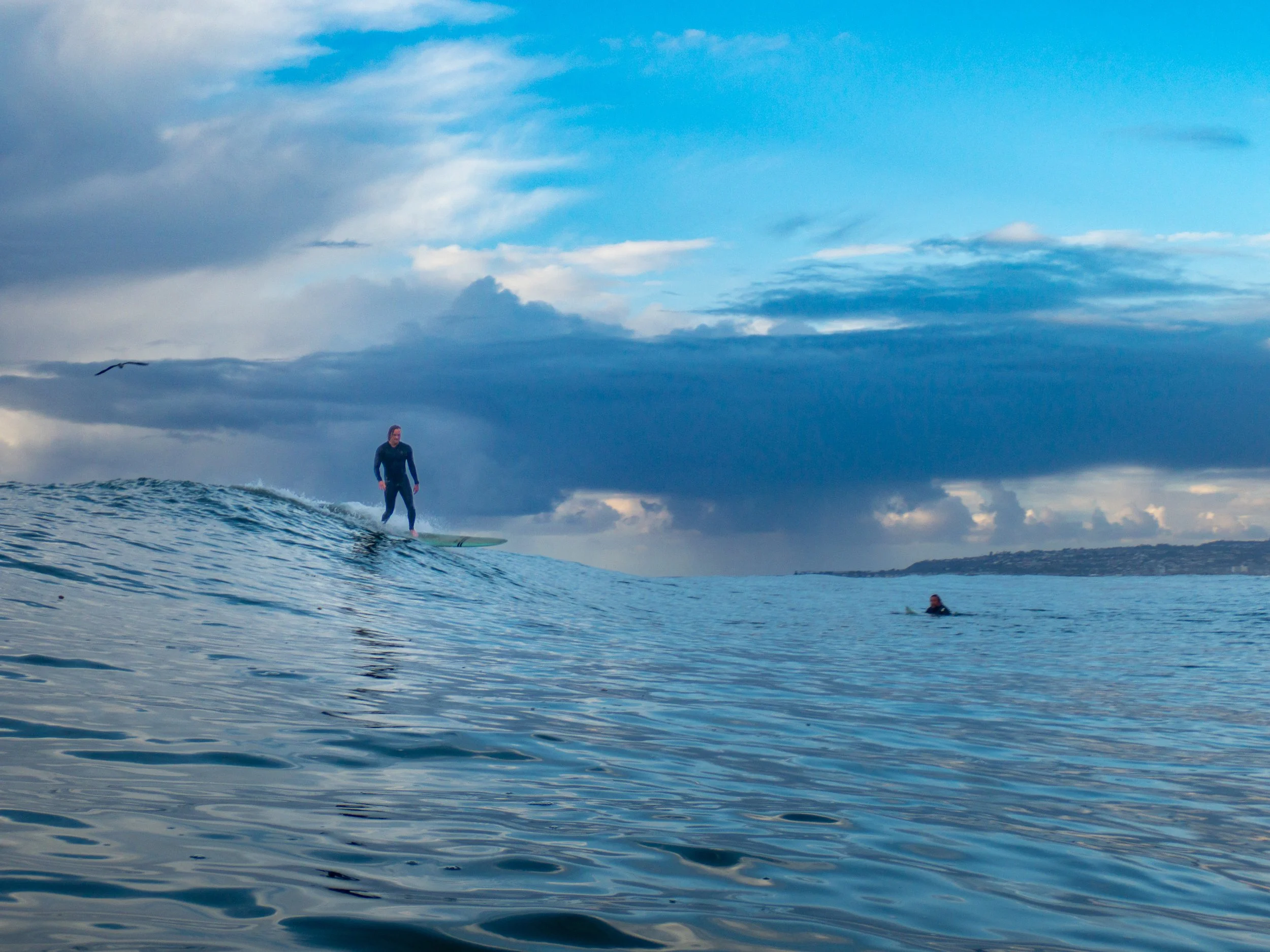 A person surfing on a wave in the ocean with another swimmer in the water nearby, under a cloudy sky.