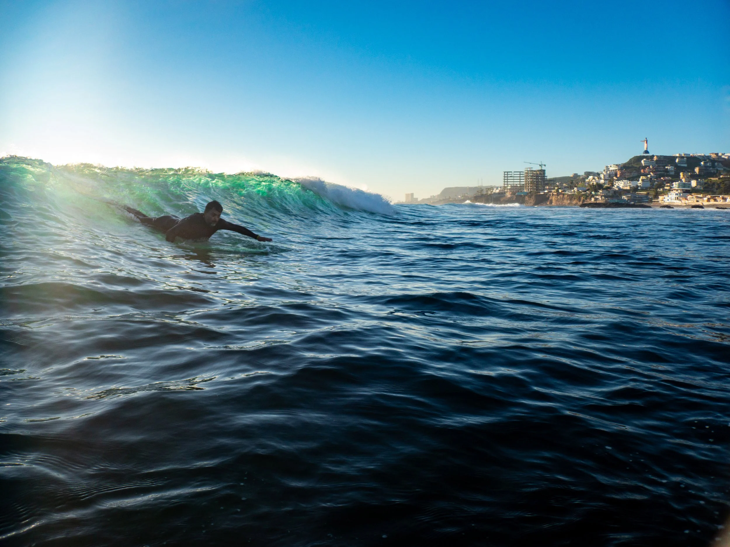 A person in a black wetsuit surfing a wave in the ocean near a coastline with buildings and a lighthouse.