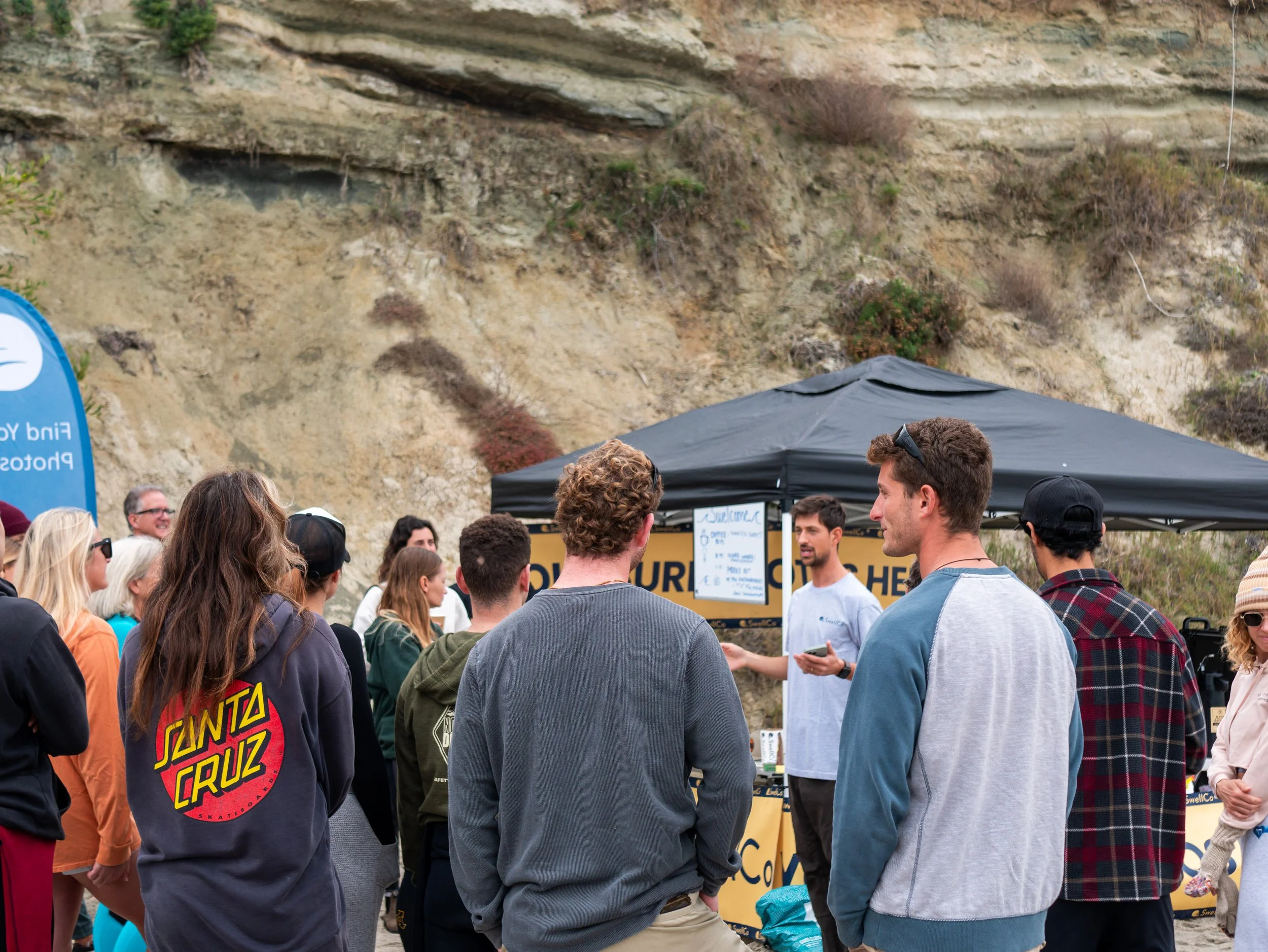 A group of people gathered outdoors under a black tent, listening to a speaker in front of a yellow sign, with rocky cliffs and sparse vegetation in the background.