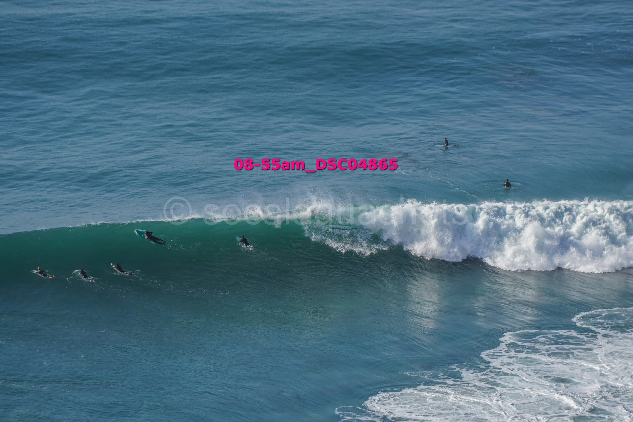 Several surfers wait for waves in the ocean, with one surfer paddling on a surfboard close to the breaking wave, and the others sitting or standing further back in the water.