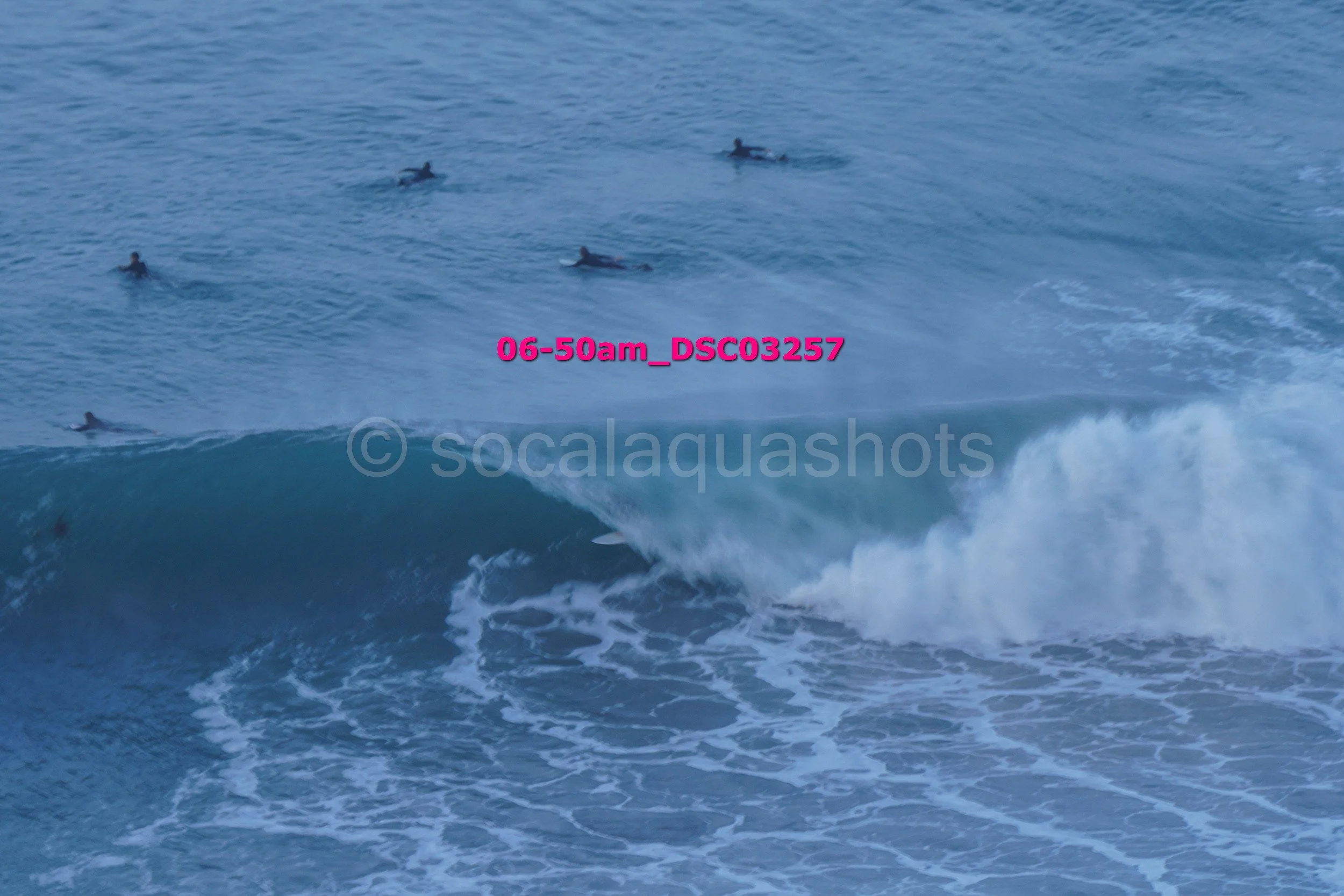 Ocean waves with several surfers in the distance, some riding and others waiting in the water.