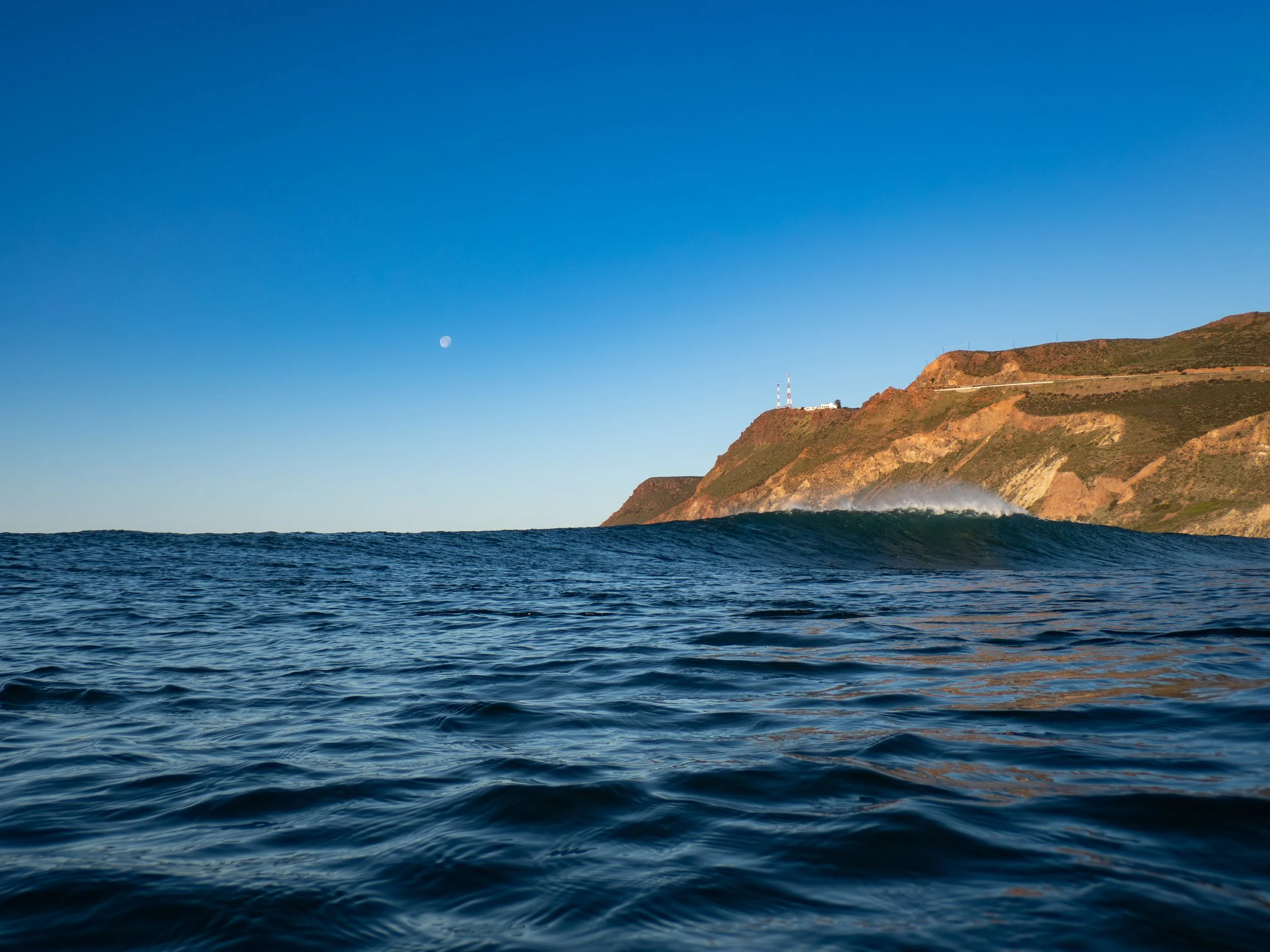 Ocean waves with a hillside and lighthouse in the distance under a clear blue sky with visible moon.