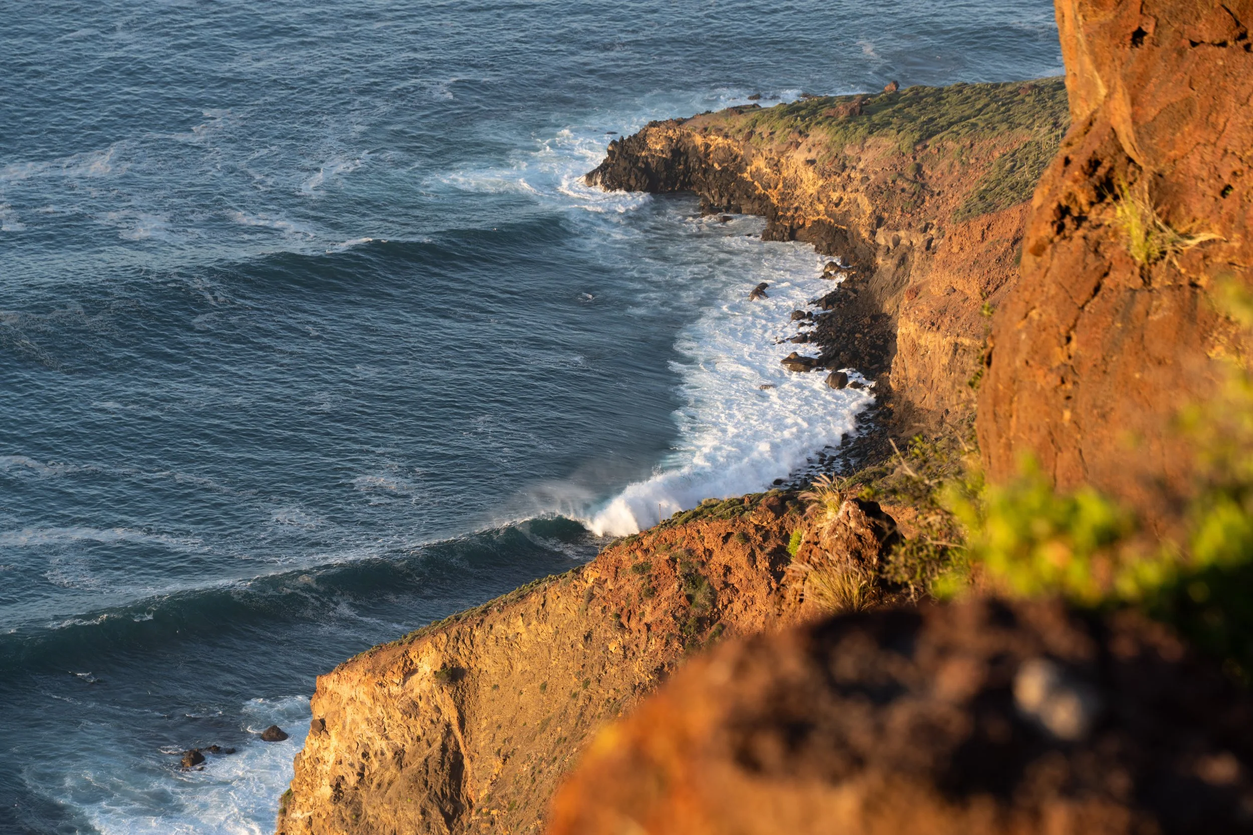 Cliffside view of the ocean with waves crashing against the rocky shoreline and sunlit reddish-brown cliffs.
